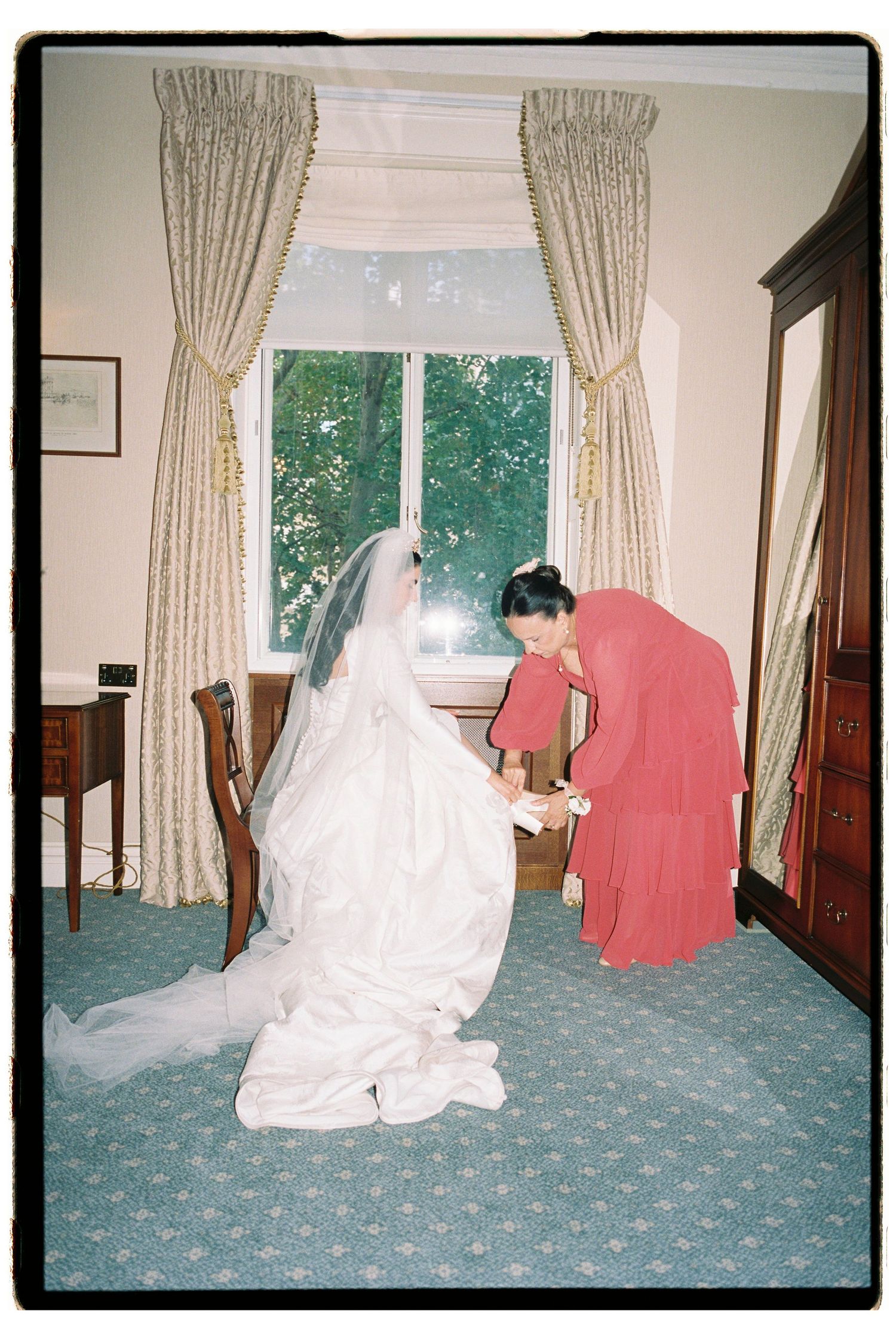 Bride in white gown and veil sits as bridesmaid in red dress assists her in an elegant room with draped curtains.