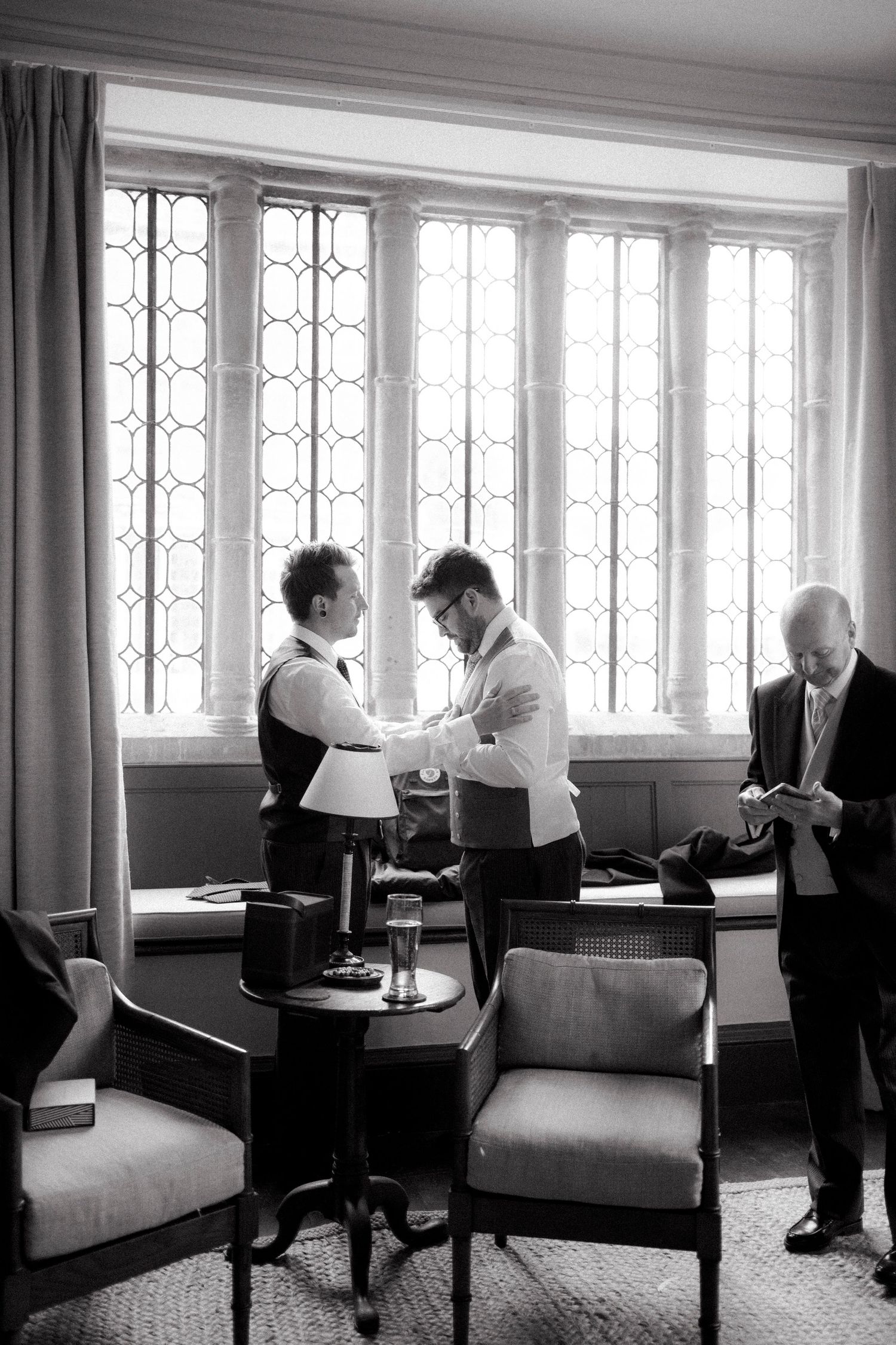 Black and white photo of groomsmen helping each other dress before a wedding ceremony near leaded glass windows.