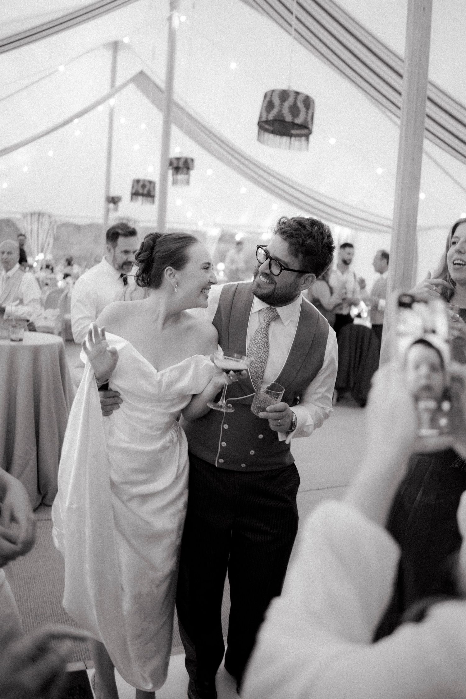 Bride and groom sharing first dance in a decorated marquee tent surrounded by wedding guests.