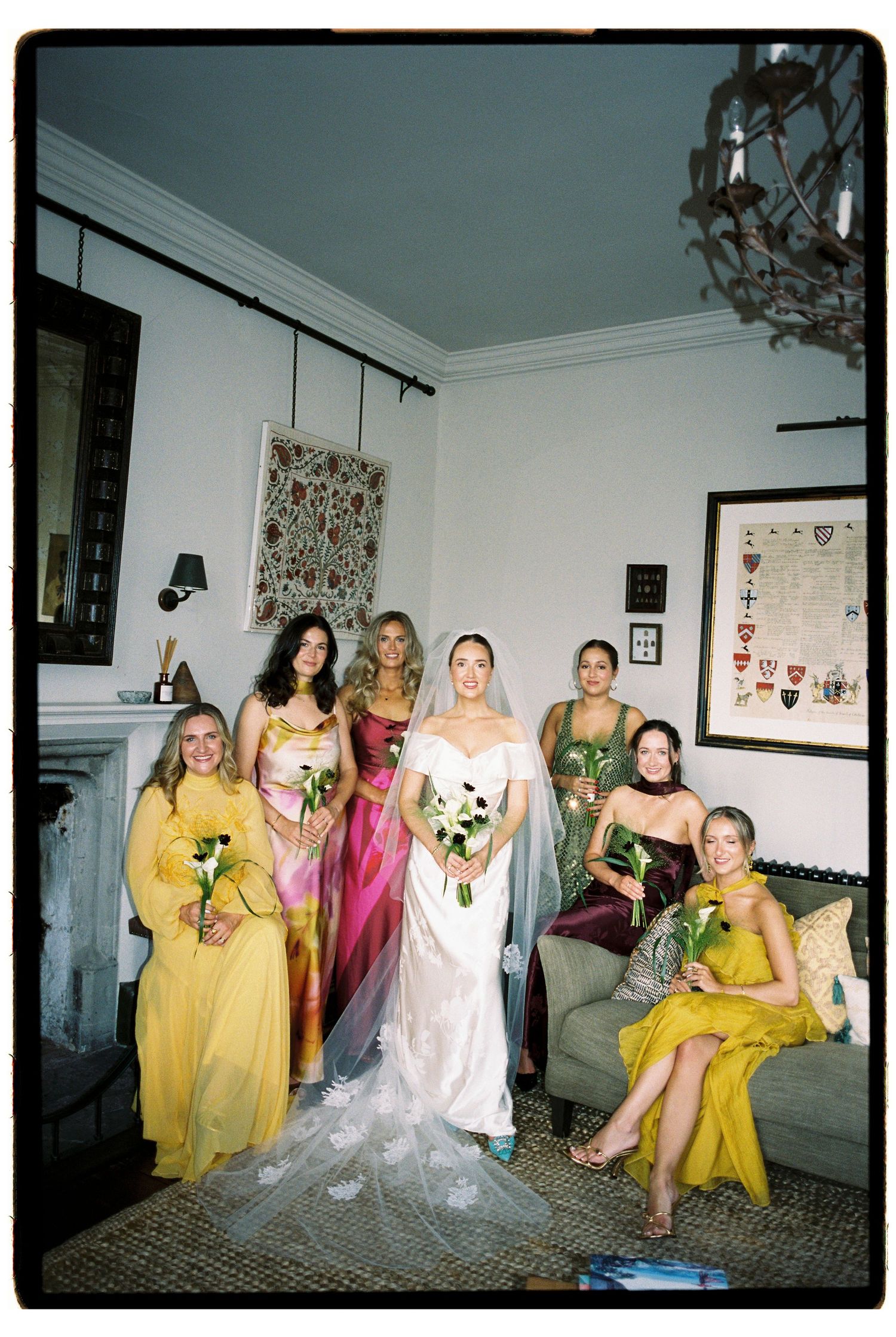 Bride in white gown poses with bridesmaids in colorful yellow, green, and pink dresses inside an elegant room.