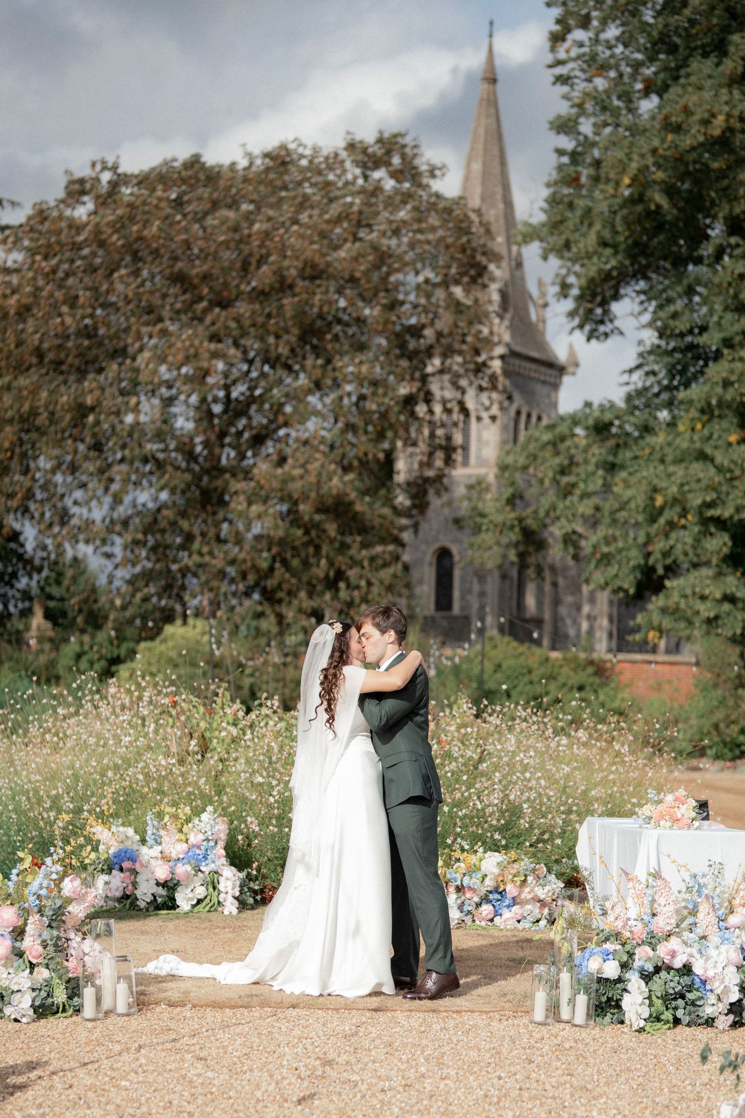 Bride and groom share first kiss at outdoor wedding with wildflowers and historic church backdrop.