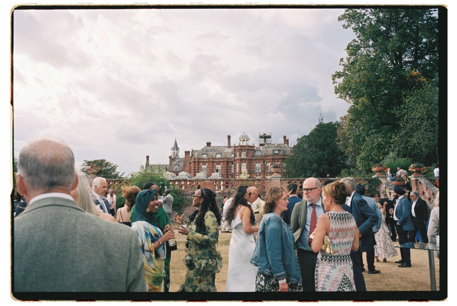 Elegant outdoor wedding reception gathering on manicured grounds of a grand English manor house on a cloudy day.