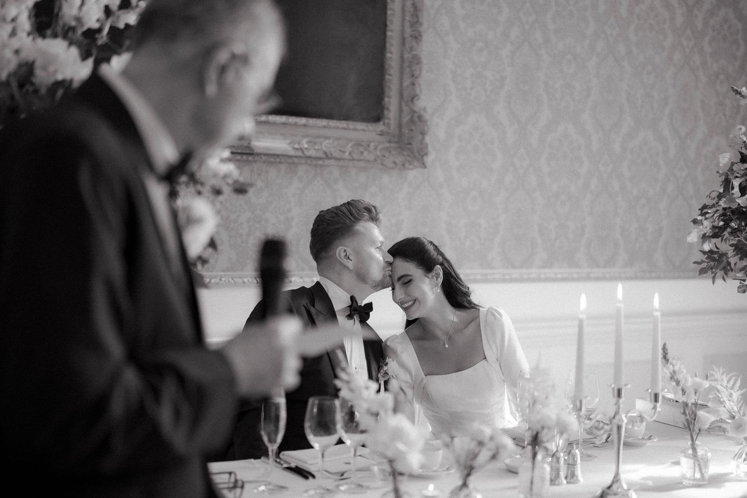 Black and white wedding photo of bride and groom sharing a tender moment during speeches at elegant reception dinner.