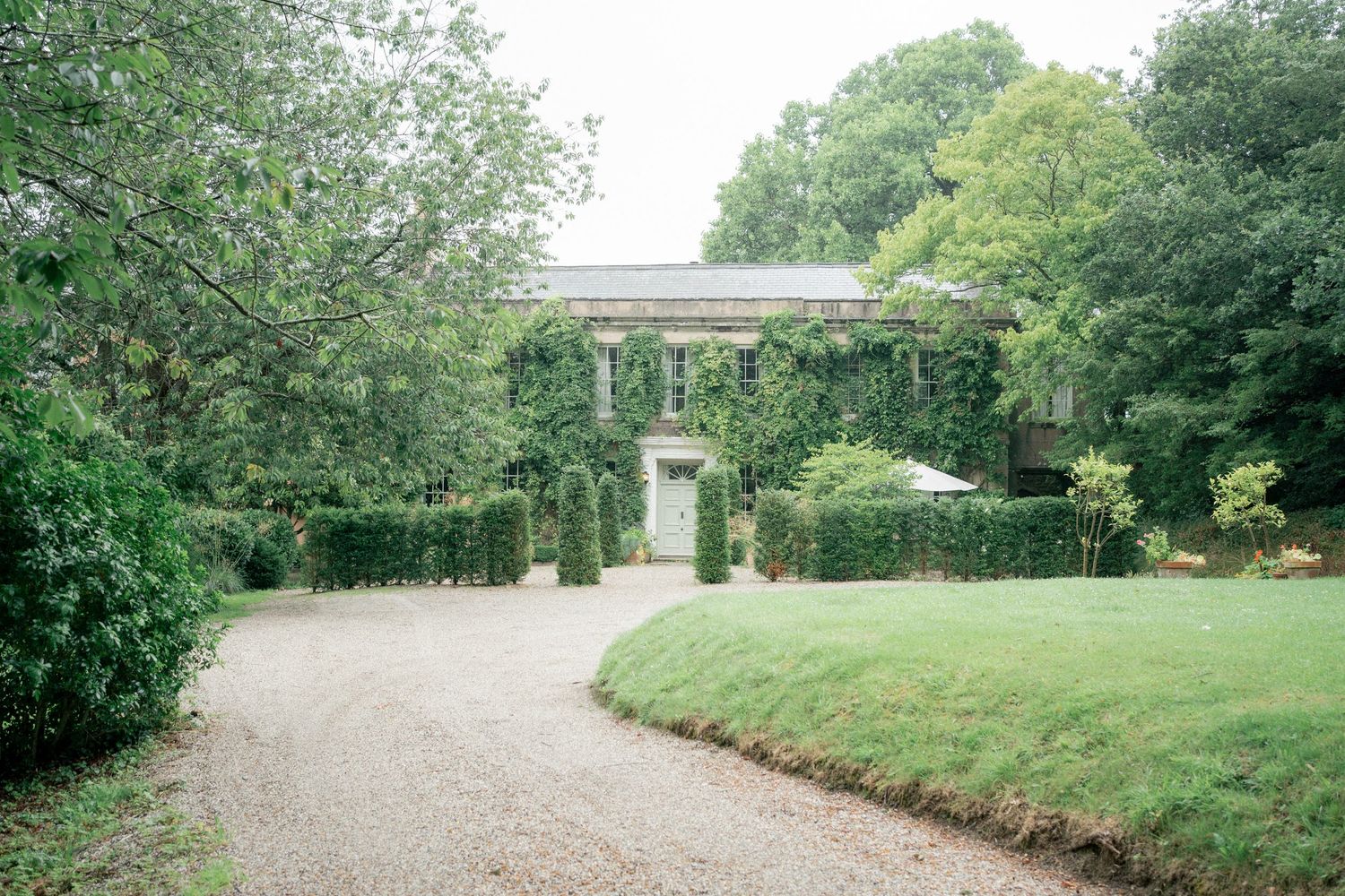 A grand country estate with ivy-covered facade, gravel driveway, manicured hedges, and lush green gardens.