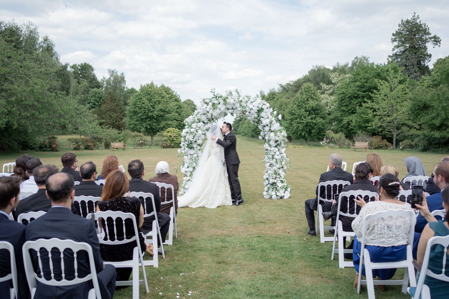 Wedding guests seated in white chairs on a grassy hill during an outdoor ceremony.