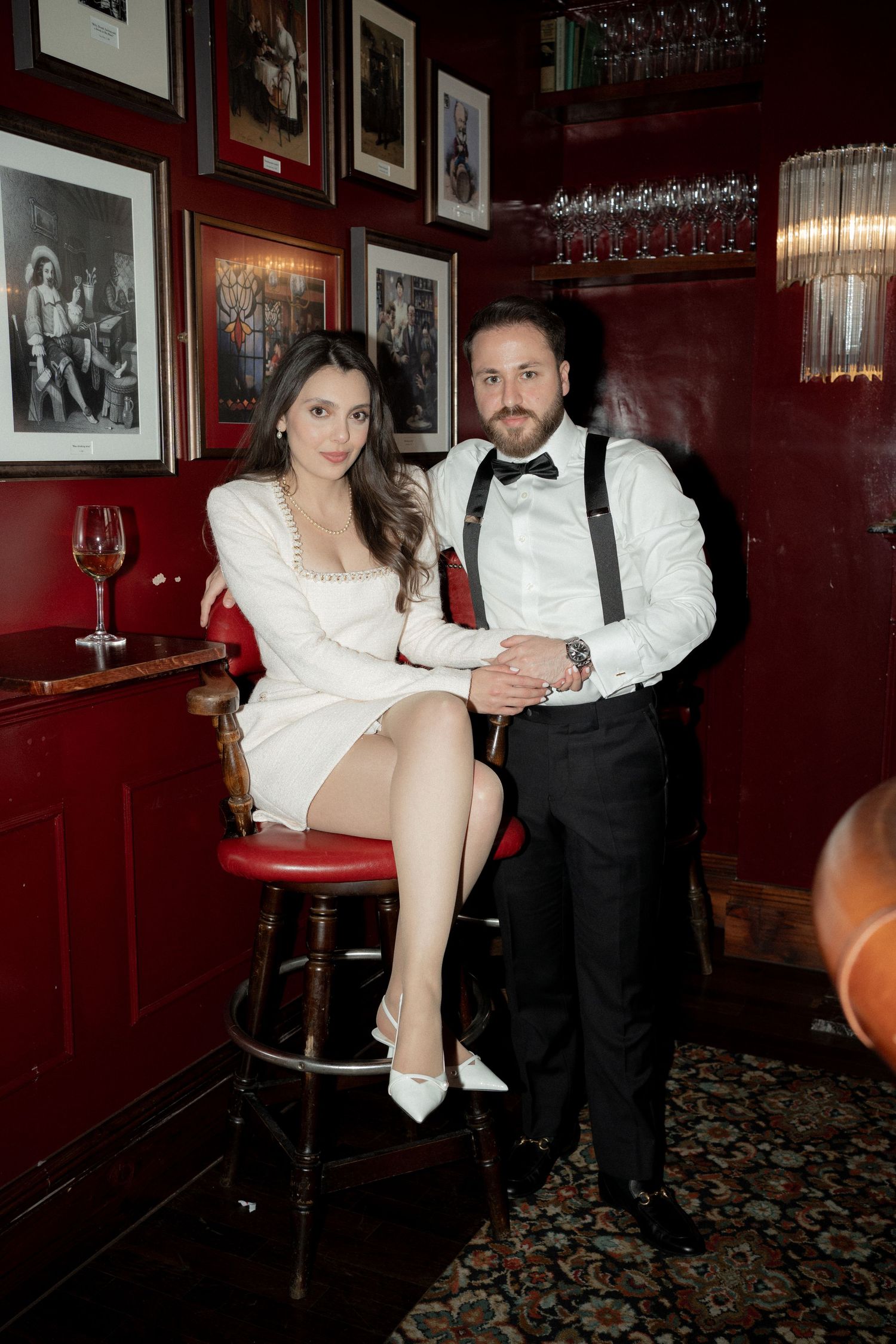 Couple in formal attire posing in a vintage-style bar with red walls and framed photographs.