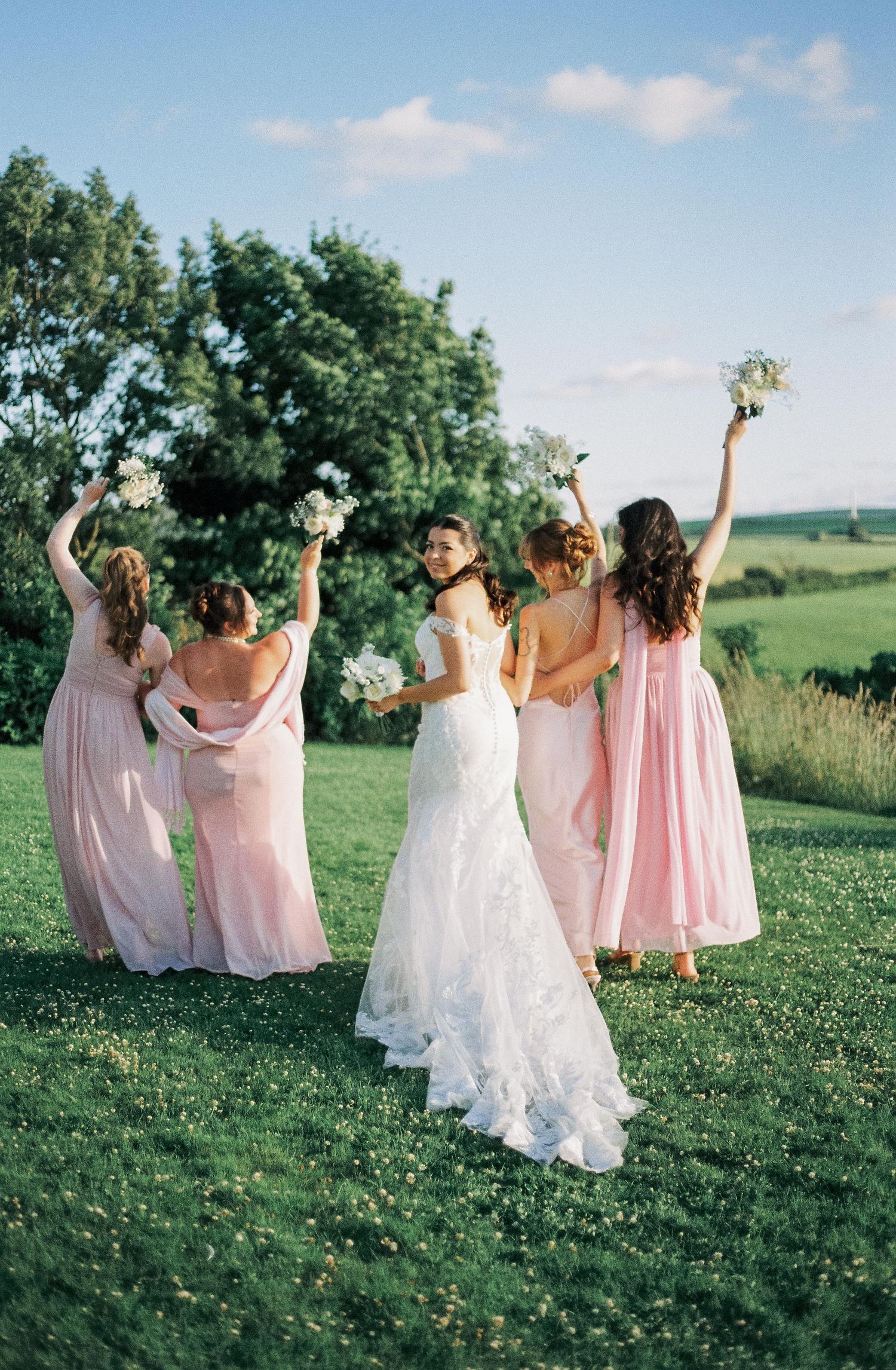 Bridesmaids in pink dresses dance joyfully on a grassy hillside during sunset.