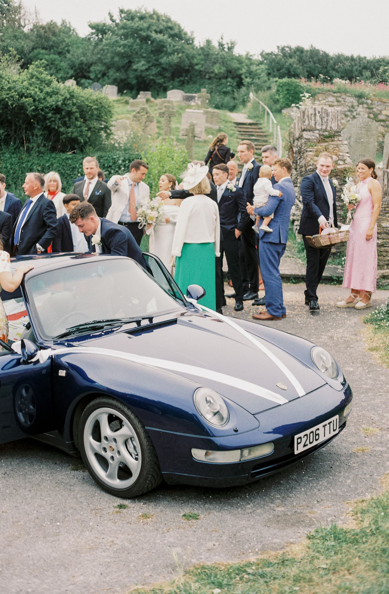 A classic navy blue Porsche sports car decorated with wedding ribbons parked at an outdoor celebration.