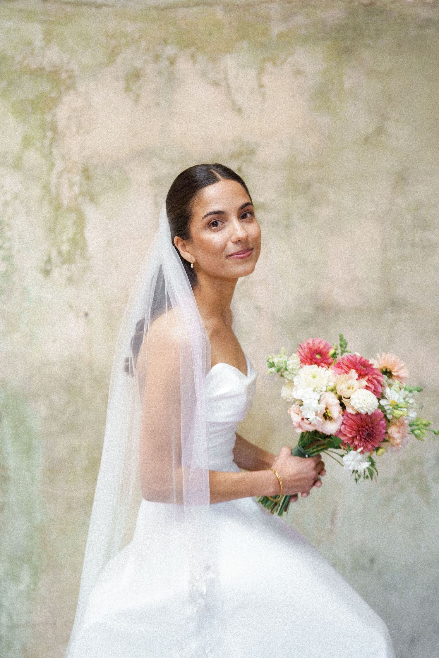 A bride in a white wedding gown holds a vibrant bouquet of pink and burgundy flowers against a textured wall.