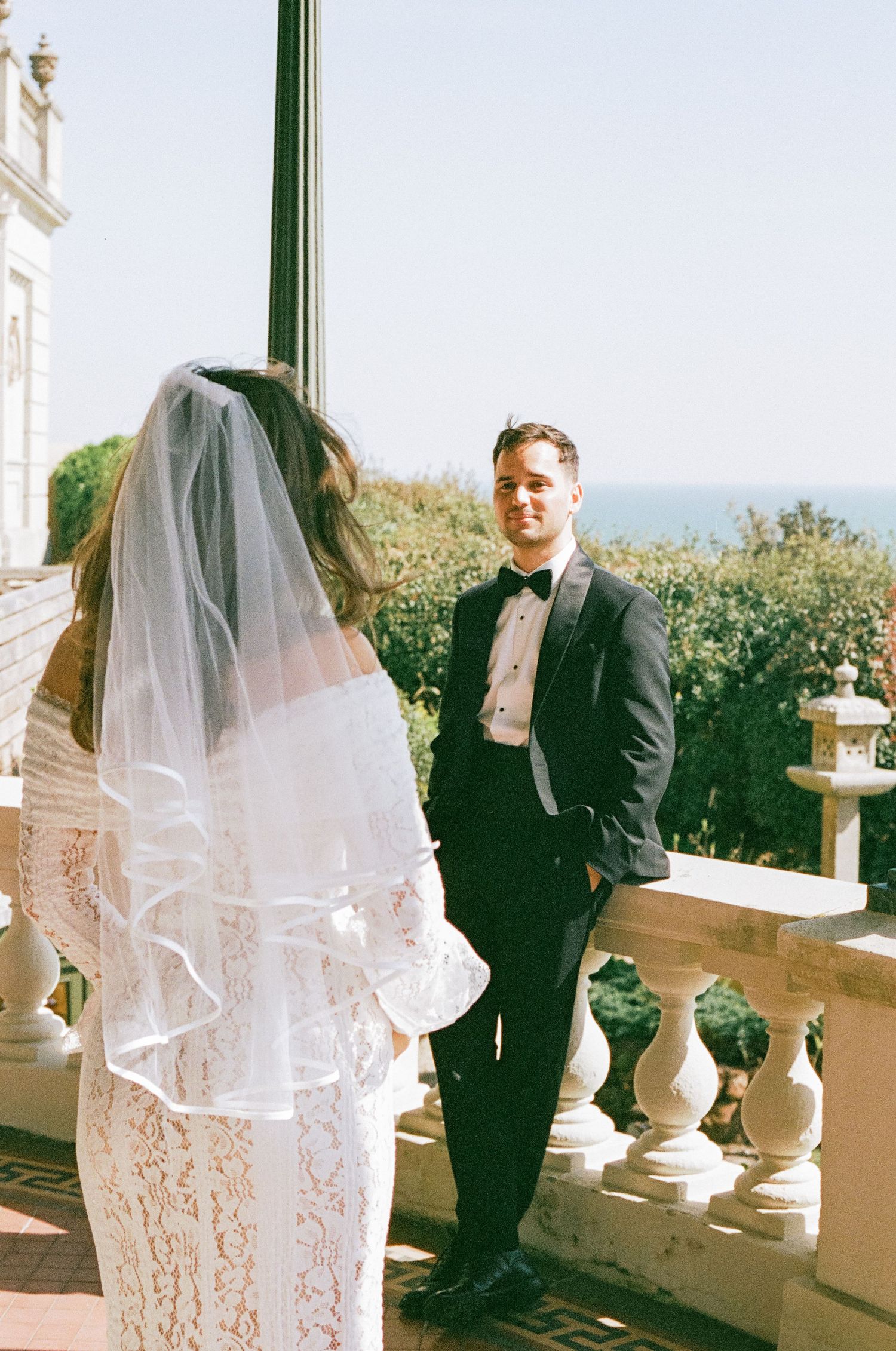 A wedding couple poses on an elegant white balcony with stone balusters overlooking an ocean view.