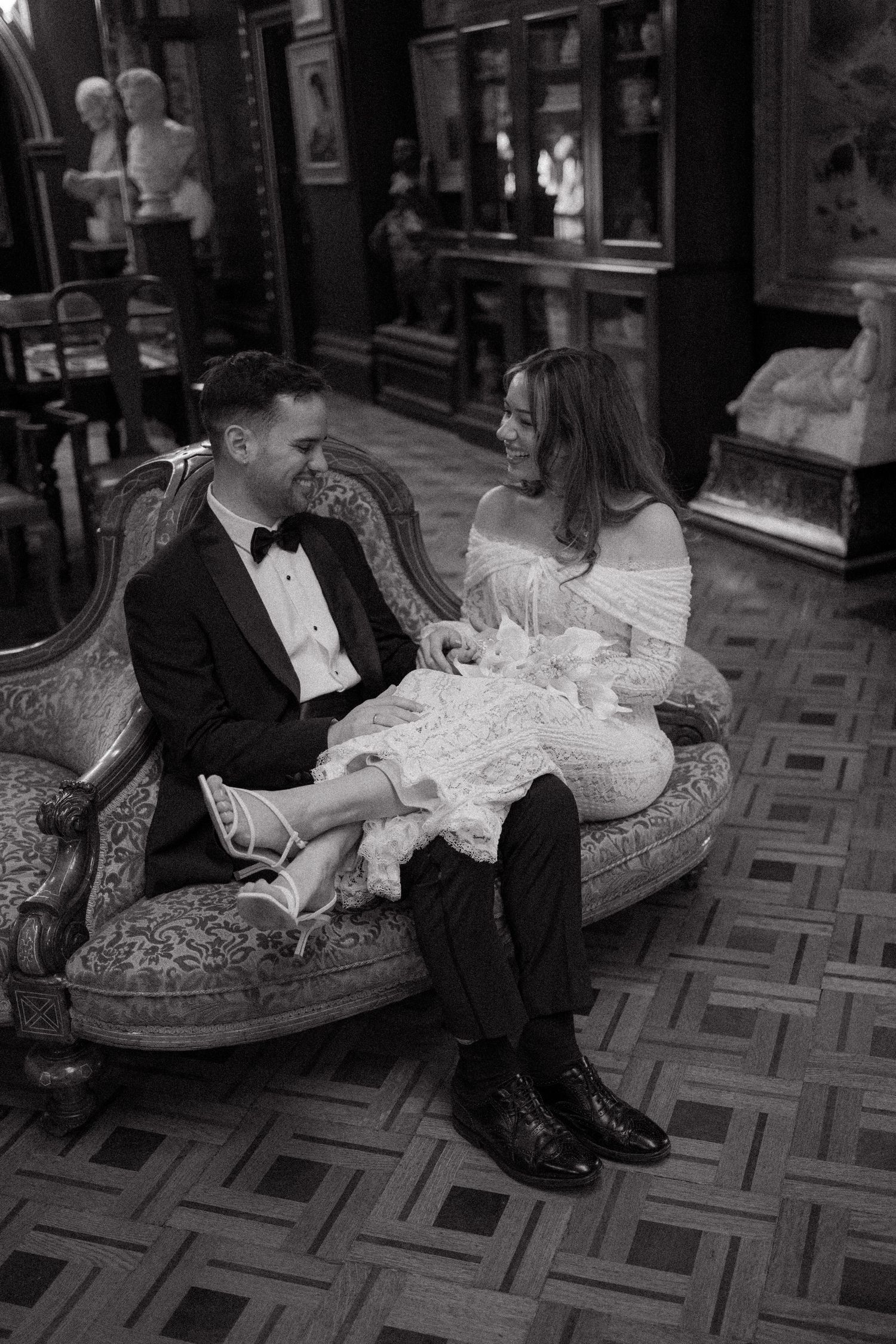 A couple in formal wedding attire sits together on an elegant patterned floor in black and white photography.