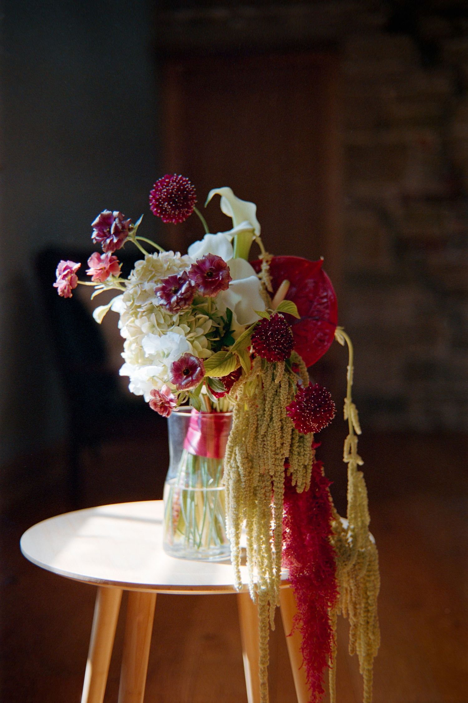 Elegant floral arrangement with cascading amaranthus and burgundy blooms displayed on a wooden side table in moody lighting.
