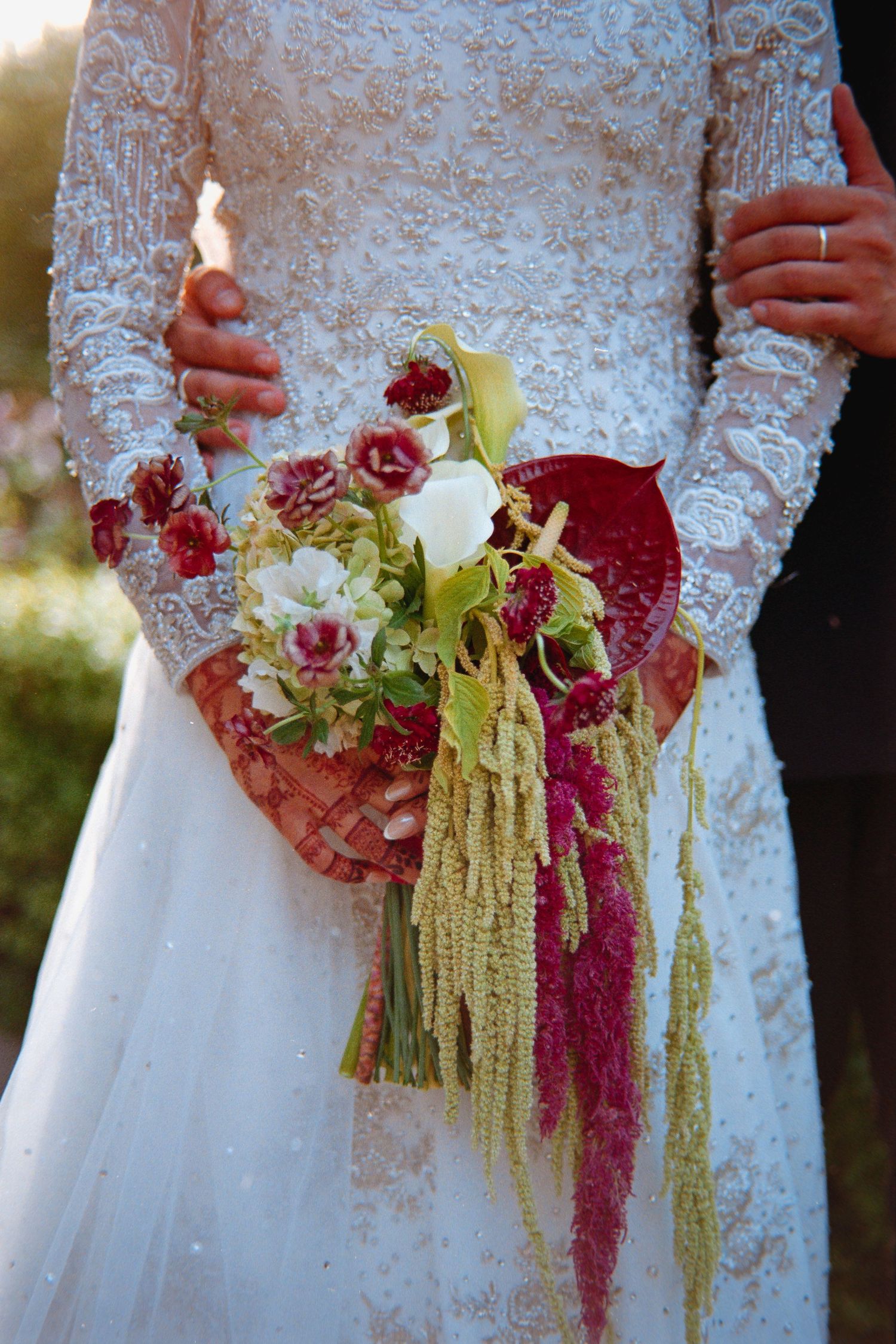 Close-up of wedding bouquets with burgundy roses and cascading amaranthus against white lace wedding dresses.