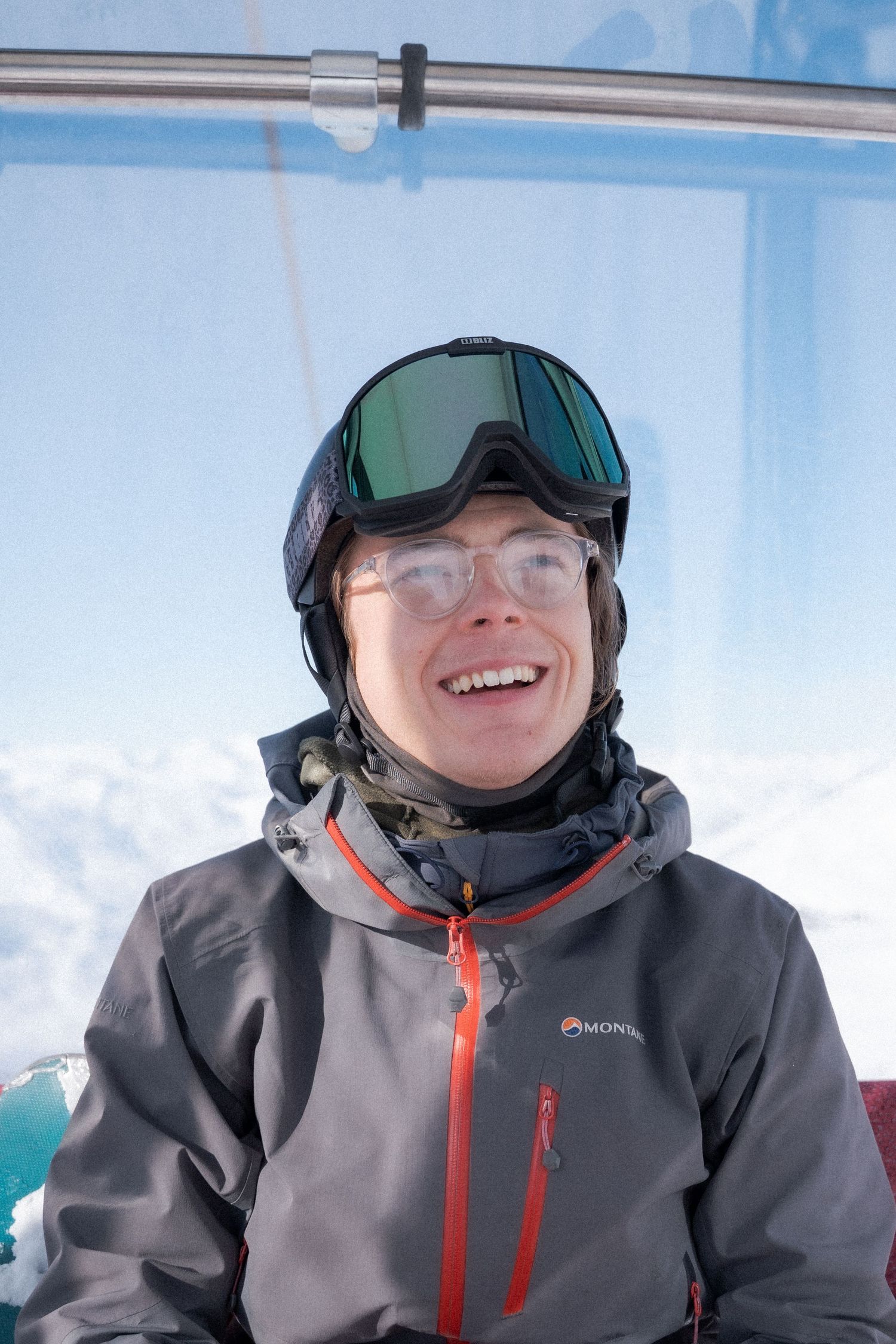 A skier wearing gray winter gear and goggles smiles while riding a chairlift on a sunny winter day.