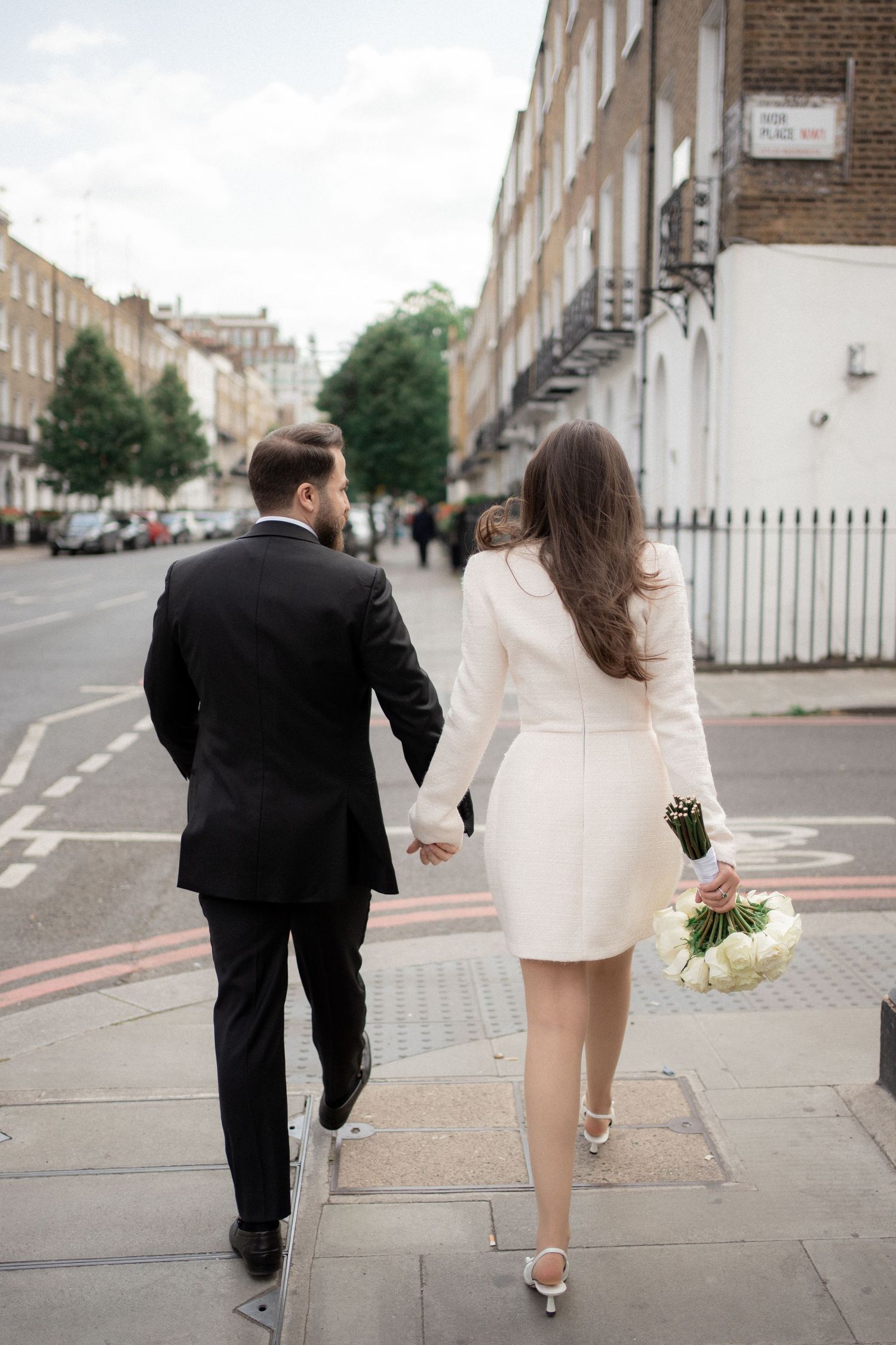 A couple in formal attire walks down a London street with the bride carrying white flowers.