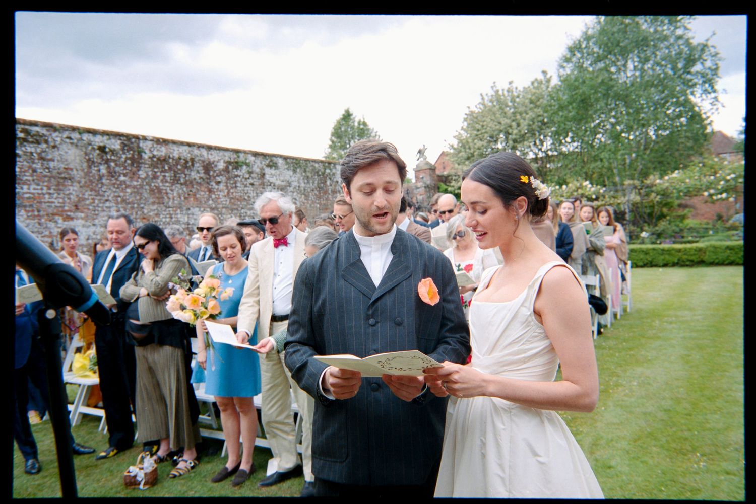 A bride and groom read their vows from paper in a garden ceremony as guests watch beside an old stone wall.