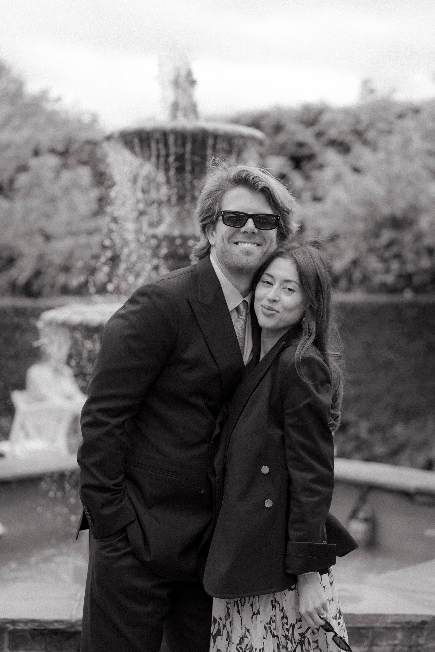 Black and white portrait of a smiling couple in dark jackets embracing in front of a stone garden fountain.