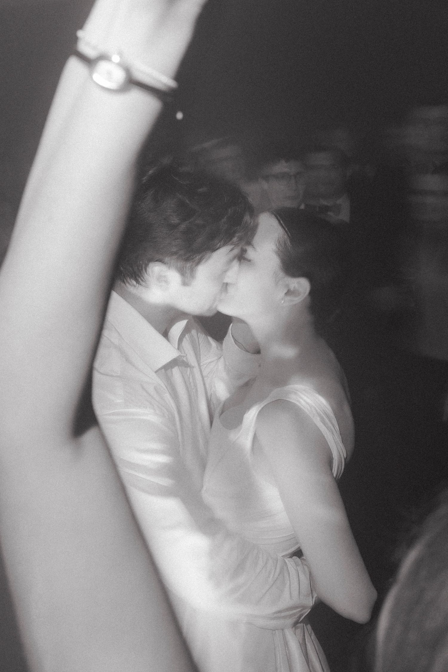 Black and white photo of a couple sharing a kiss on the dance floor at a wedding reception while a guest raises an arm in the foreground.