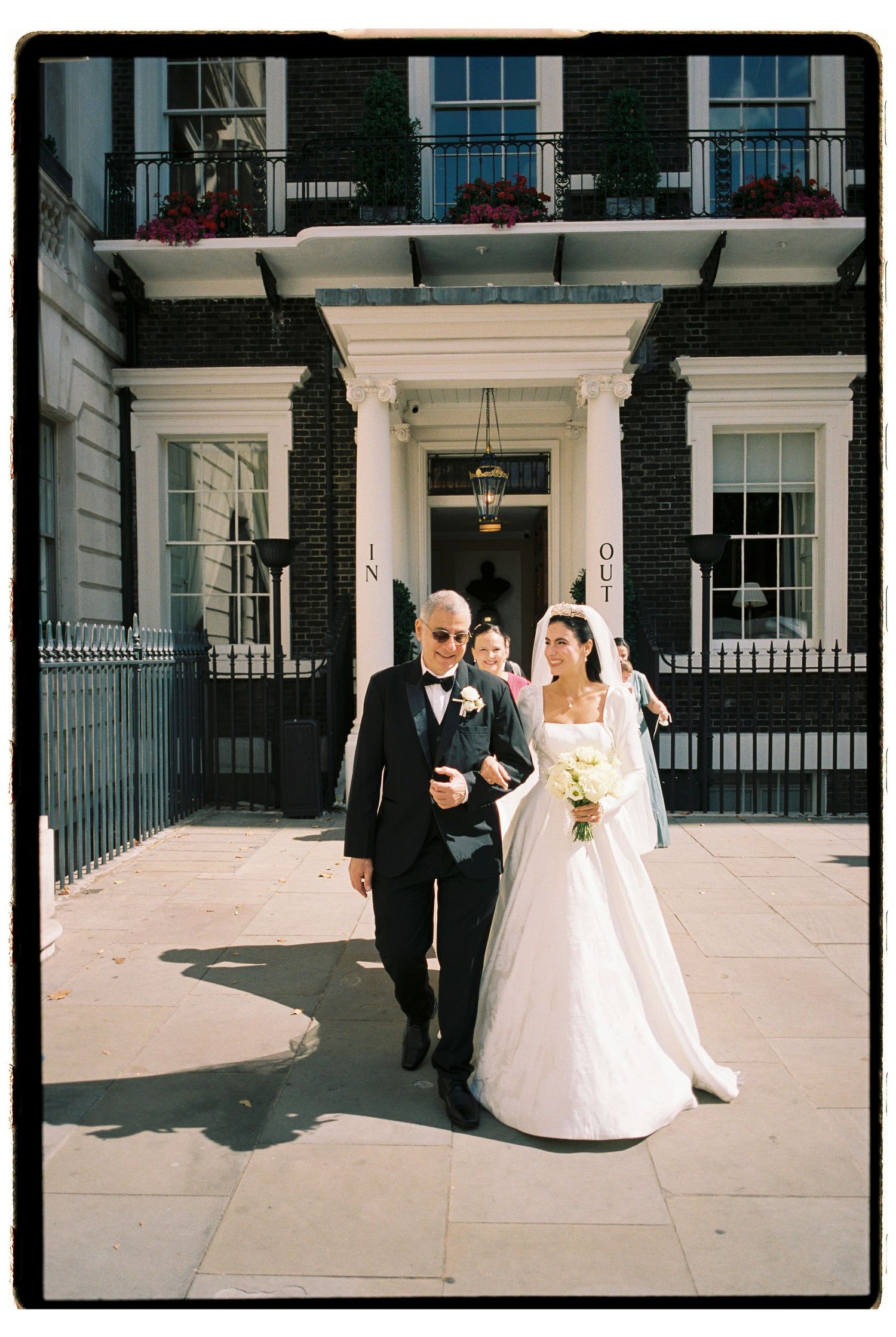 Bride in white gown walks with older gentleman in tuxedo outside elegant Georgian building on sunny wedding day.