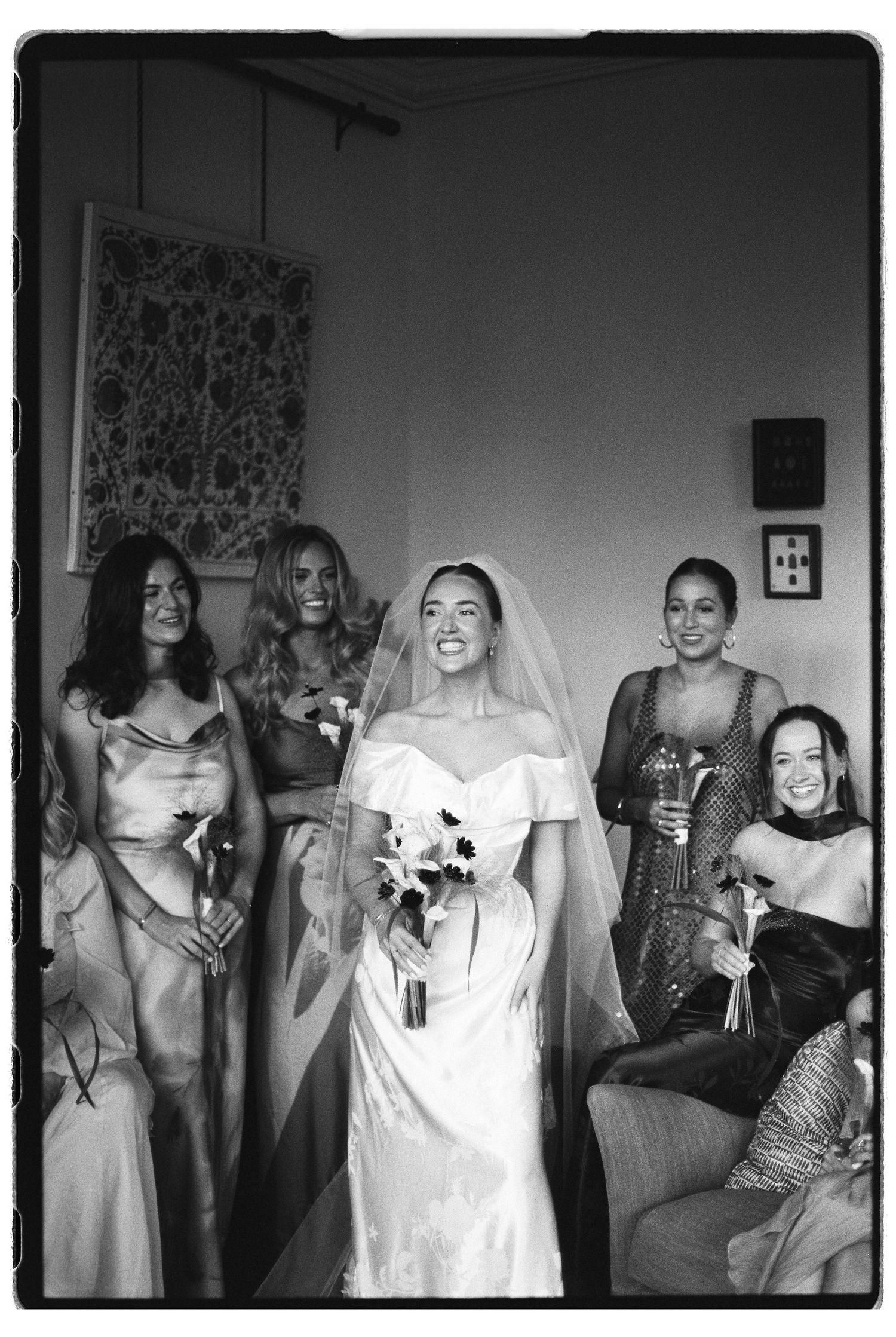 Bride in white gown and veil holds bouquet surrounded by smiling bridesmaids in elegant dresses, black and white film photo.