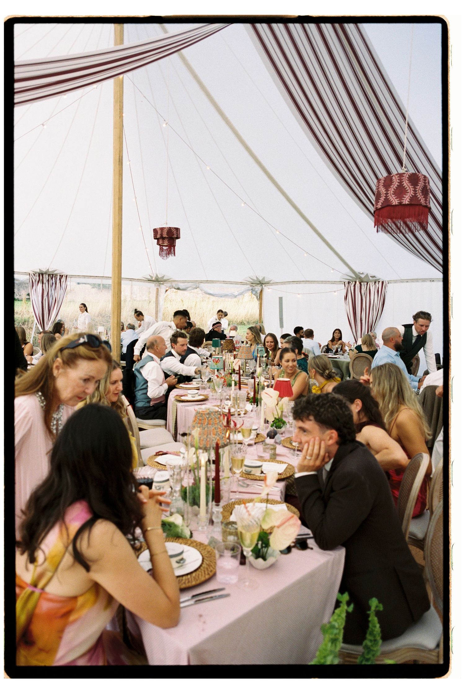 Guests mingle and dine under a decorated tent at an elegant outdoor wedding reception with pink table settings.
