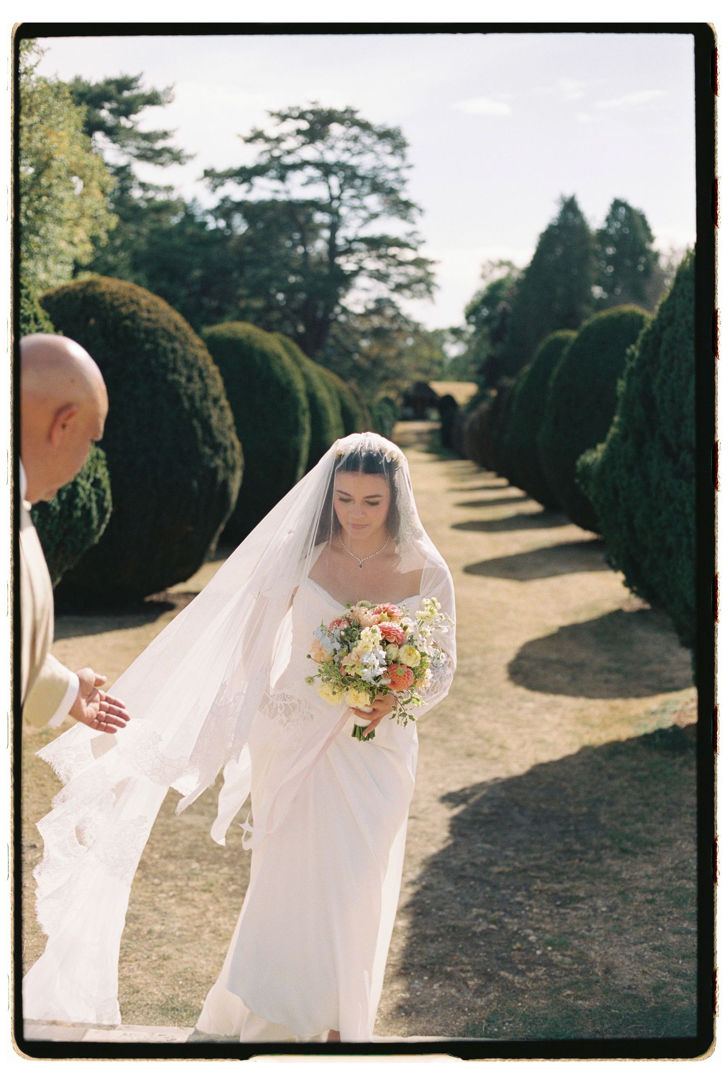 Bride in white gown and veil walks through manicured topiary garden holding colorful bouquet on wedding day.