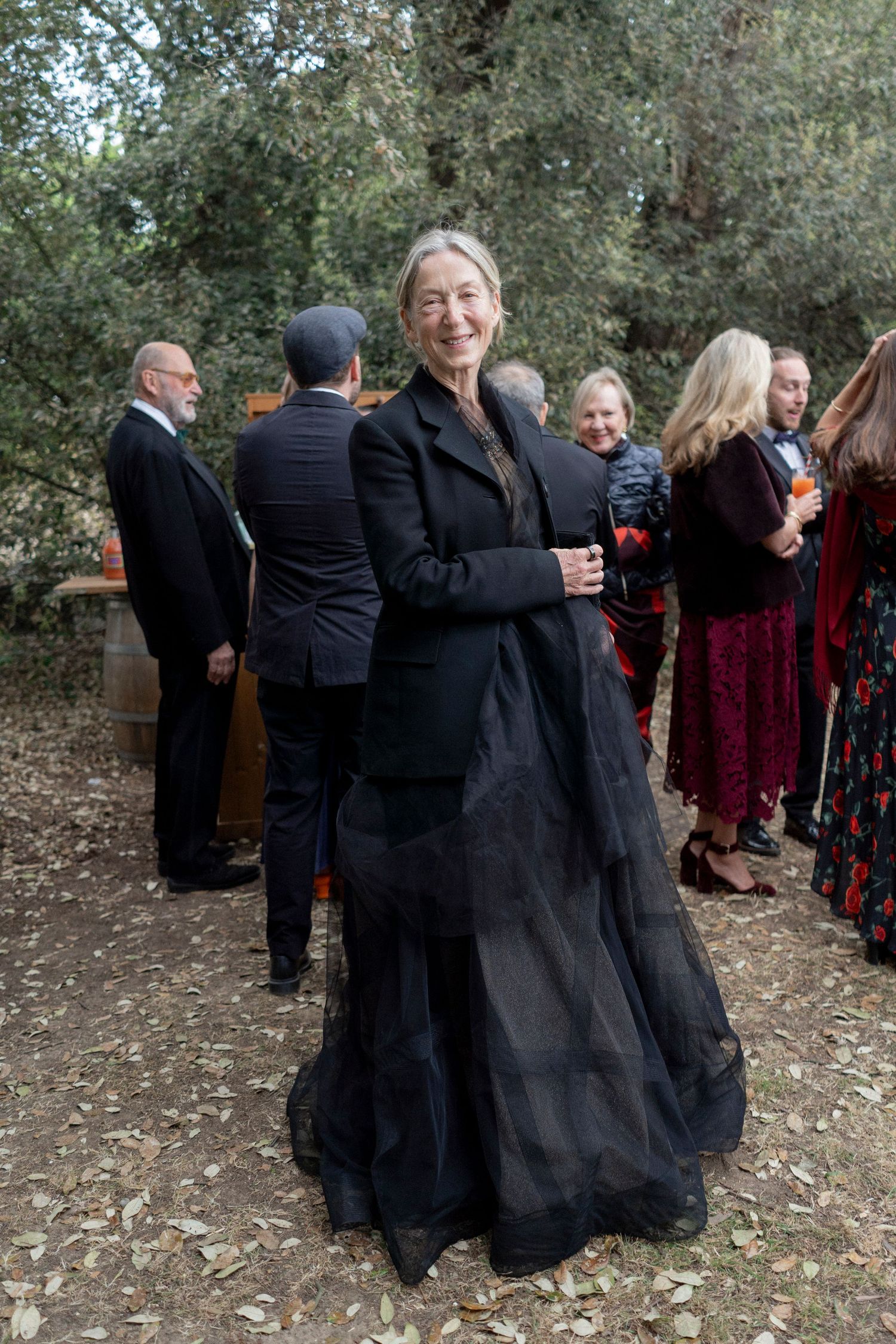 Smiling woman in a black jacket and flowing tulle skirt stands on a leaf-covered path at an outdoor gathering under trees.