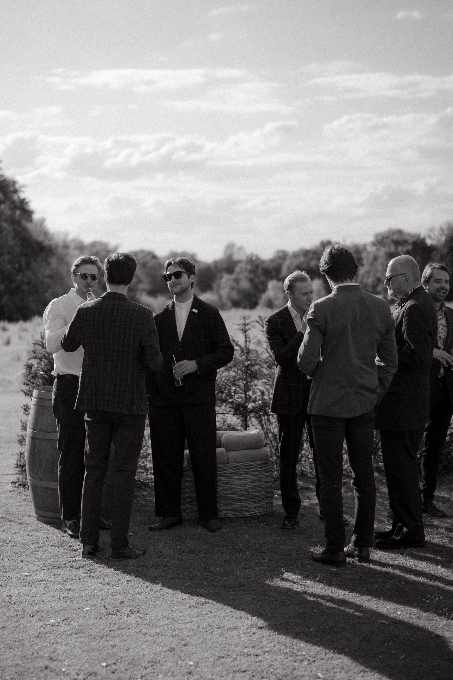 Black and white photo of wedding guests in suits chatting outdoors in a sunlit field with trees and a cloudy sky.