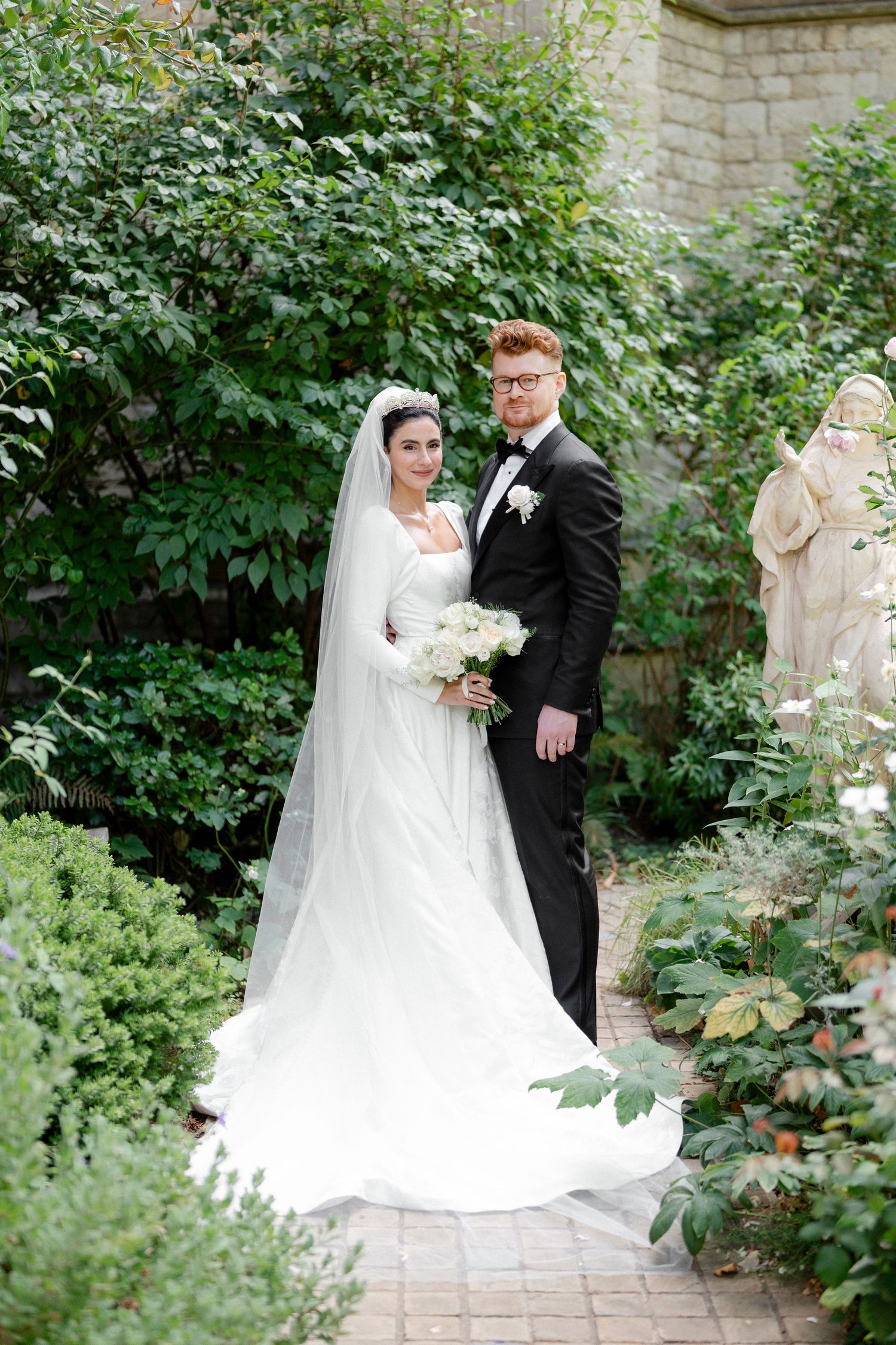 Bride and groom posing in lush garden, bride wearing white gown with veil holding white bouquet.