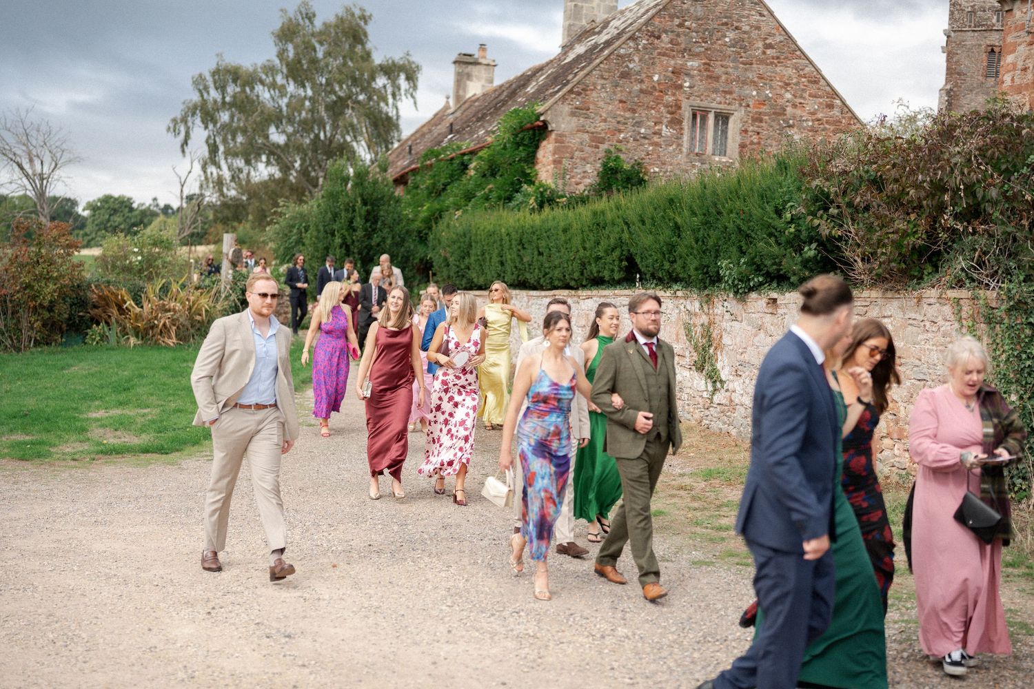 Wedding guests in colorful attire walk along a gravel path past a stone building during an outdoor celebration.