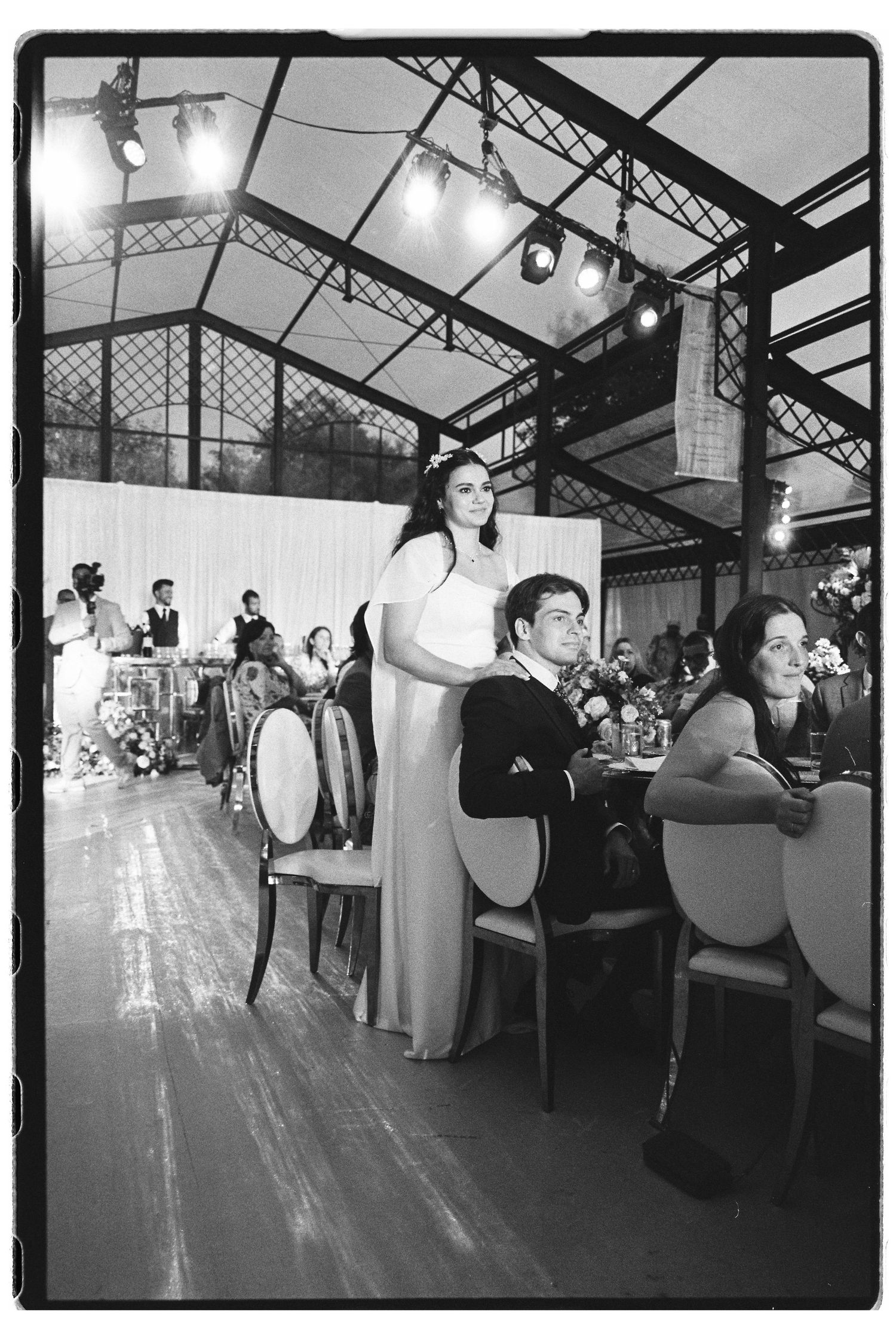 Bride and groom stand at the head table during their wedding reception in an elegant glass-roofed venue.
