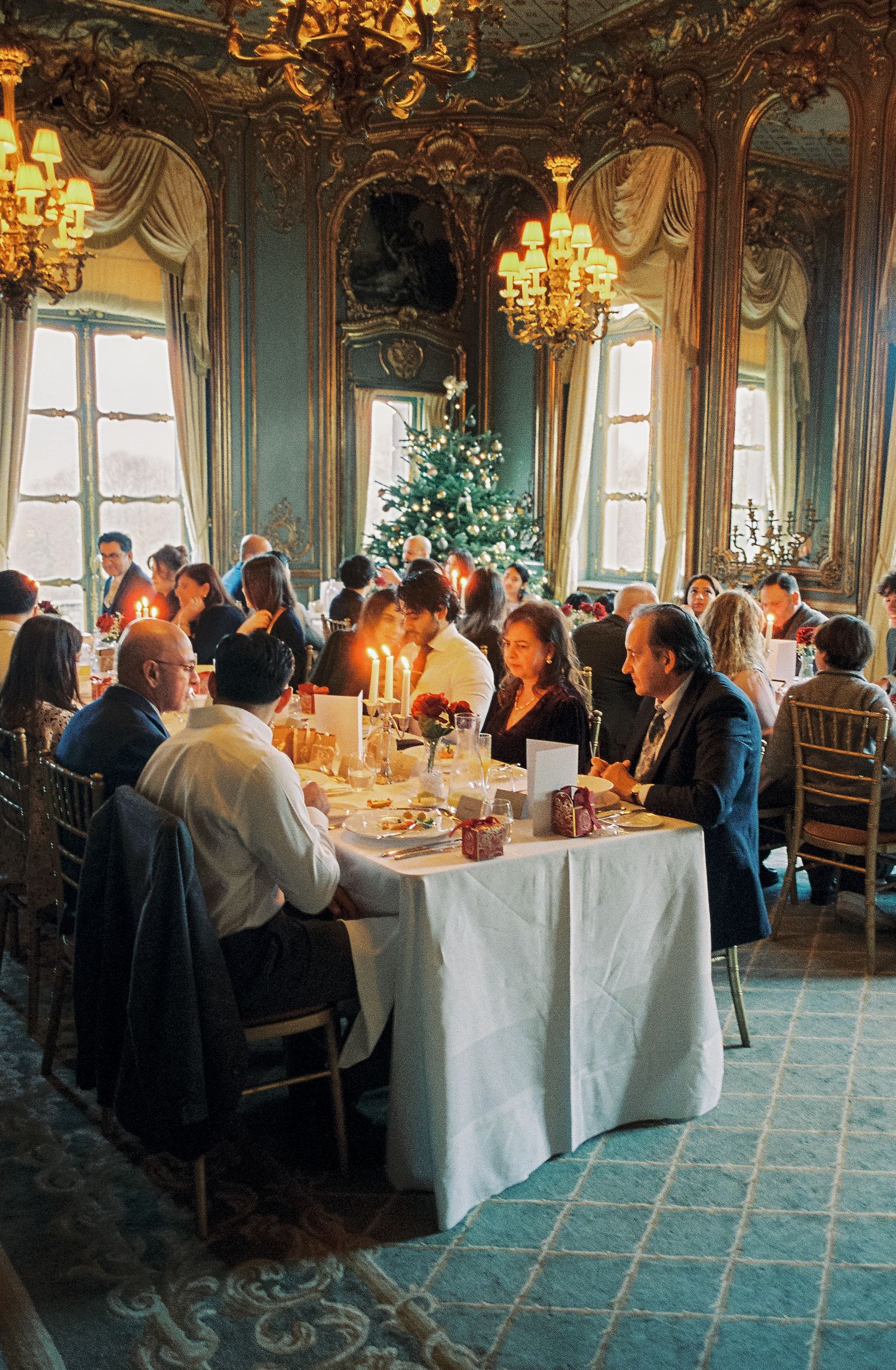 Elegant Christmas dinner party in ornate baroque dining room with candlelit tables, chandeliers, and decorated holiday tree.