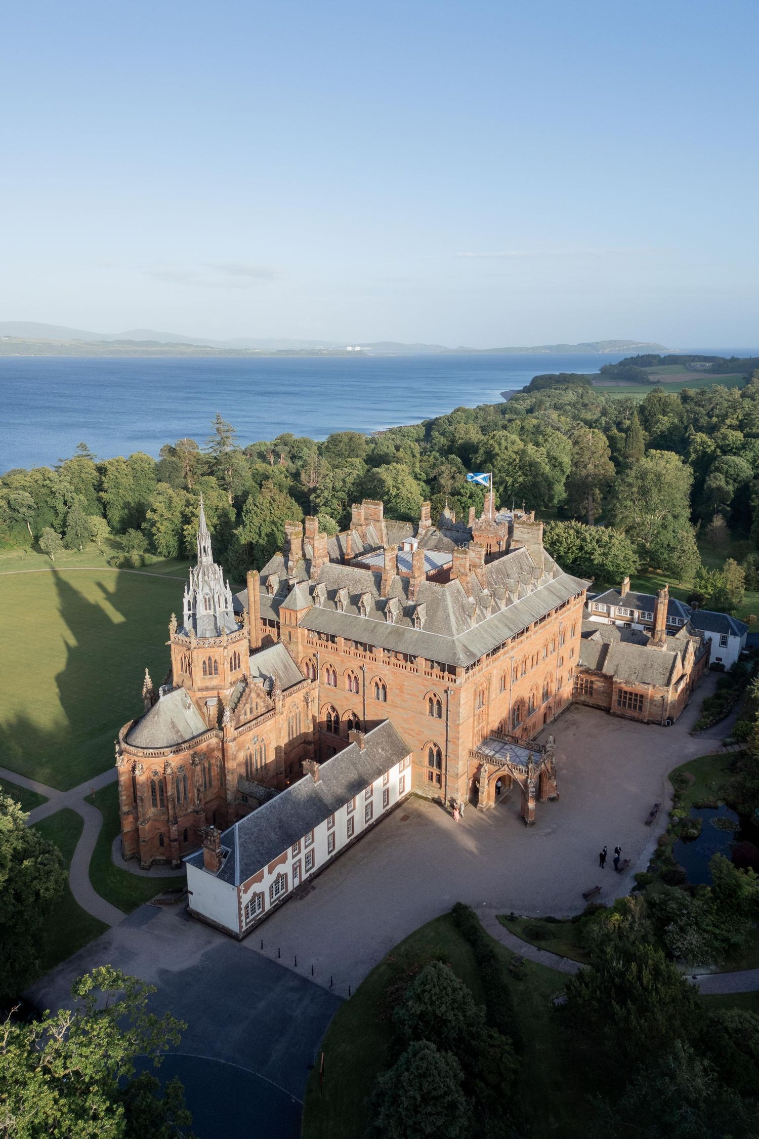 Aerial view of a grand historic mansion with red brick architecture surrounded by lush green grounds on a sunny day.