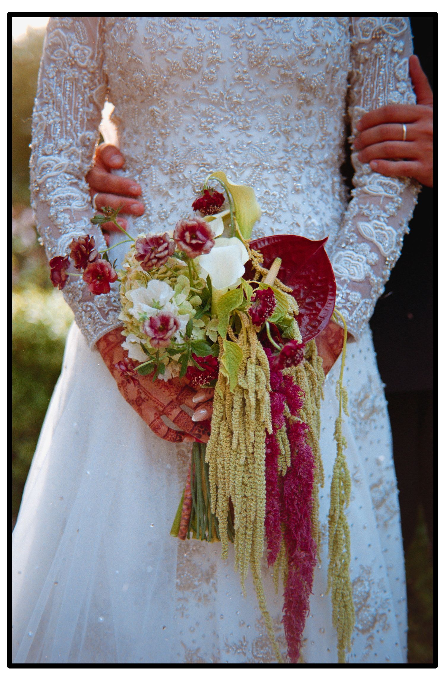 A cascading bridal bouquet features burgundy orchids and trailing amaranthus against a white lace wedding dress.