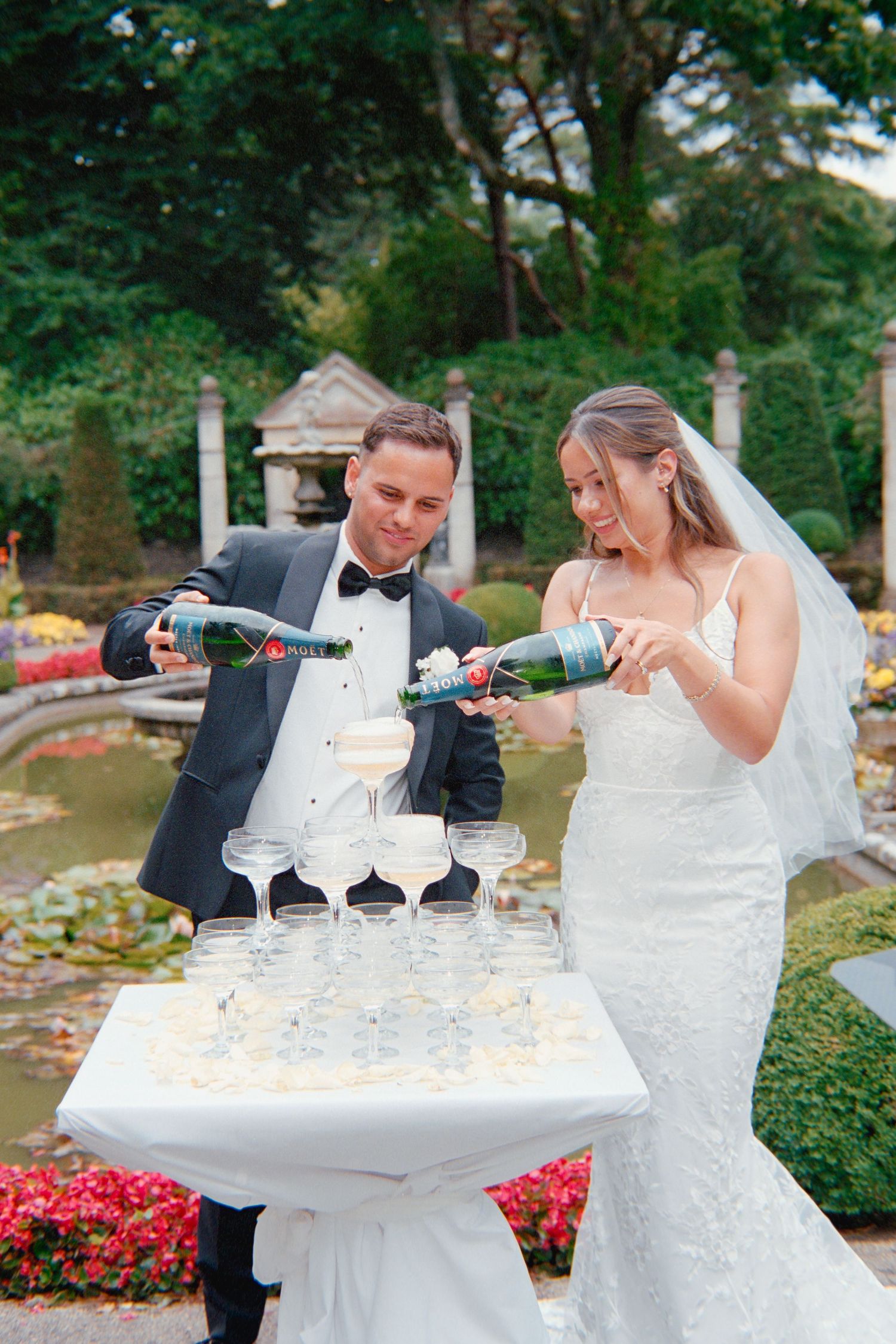 Couple pouring champagne into a tower of glasses at an elegant outdoor garden wedding celebration.