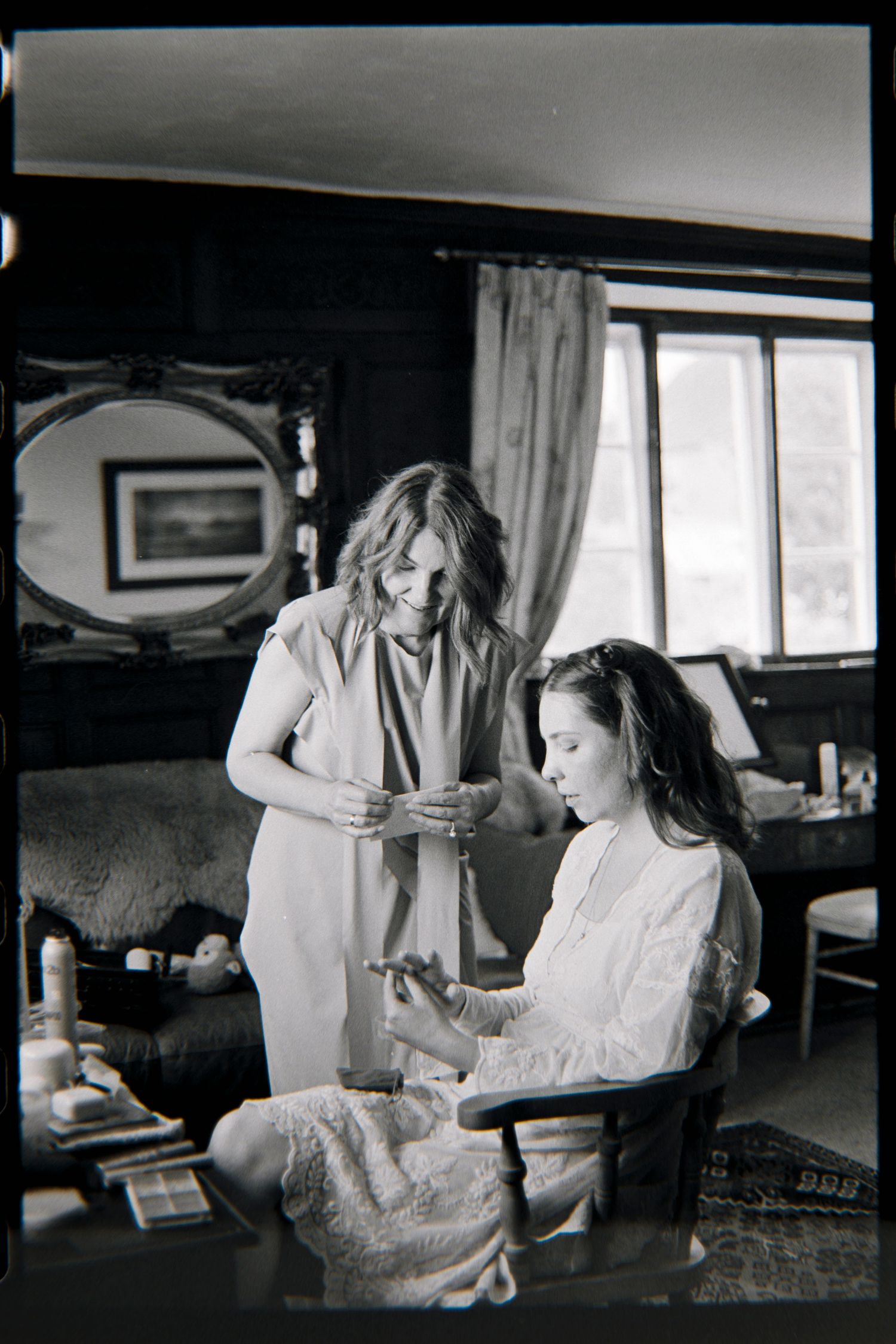 Black and white photo of a woman standing beside a seated woman in a lace dress as they prepare in a cozy room with a mirror.