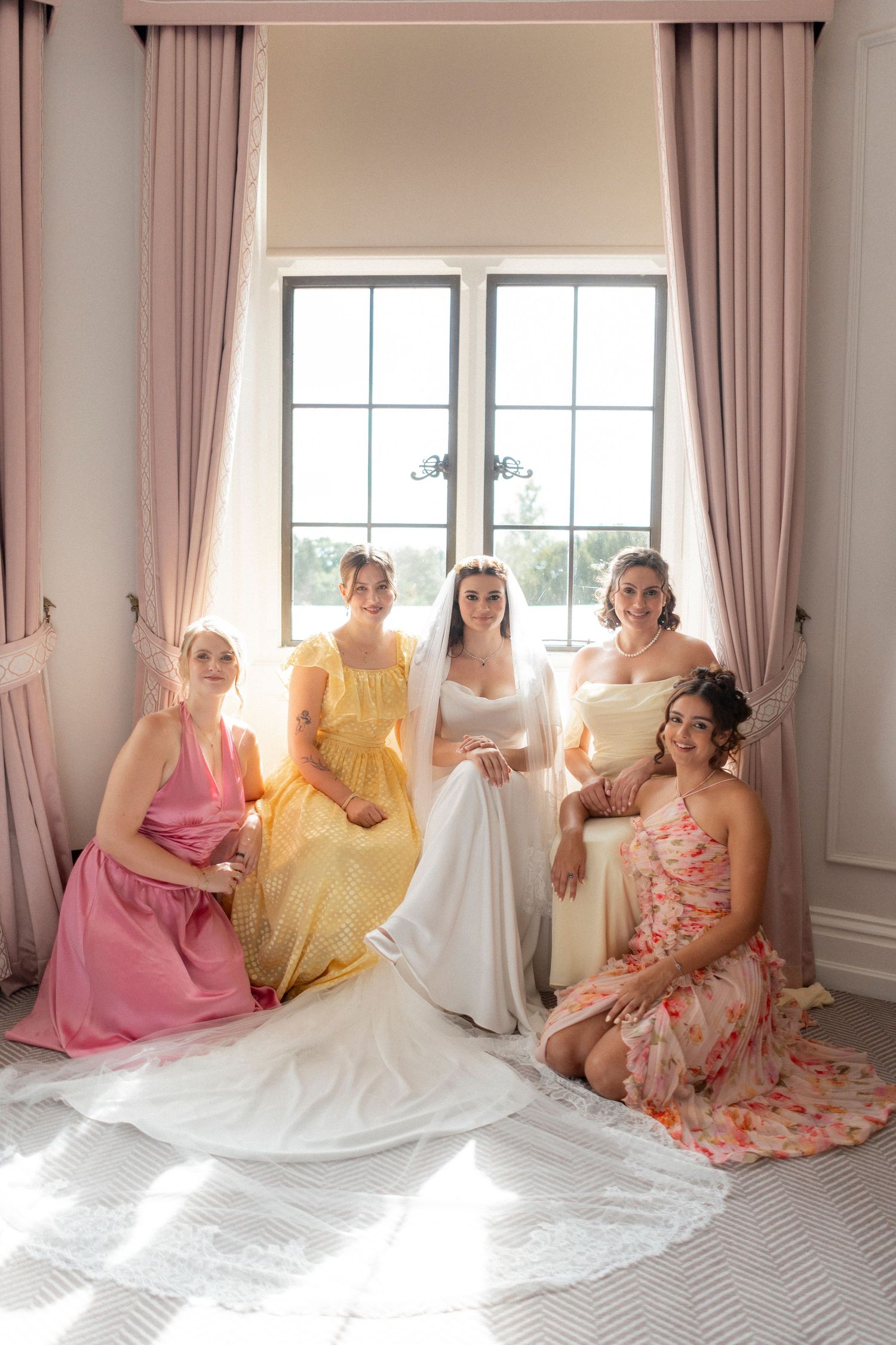 A bride in white gown poses with four bridesmaids in colorful dresses by a sunlit window with pink curtains.