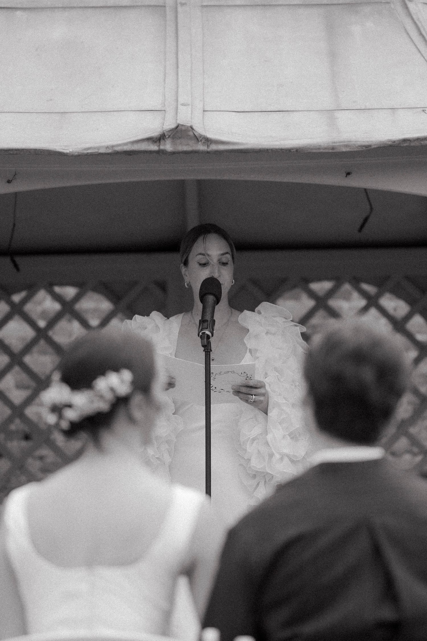 Black and white photo of a bride reading a speech into a microphone under a gazebo as wedding guests listen in the foreground.