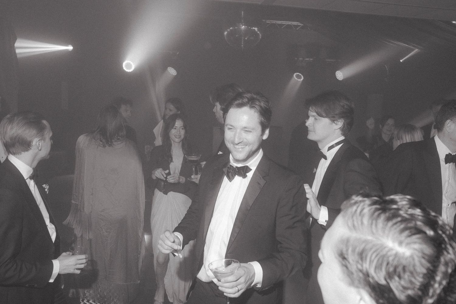 A man in a tuxedo and bow tie smiles while holding a drink among formal guests at an elegant evening party.