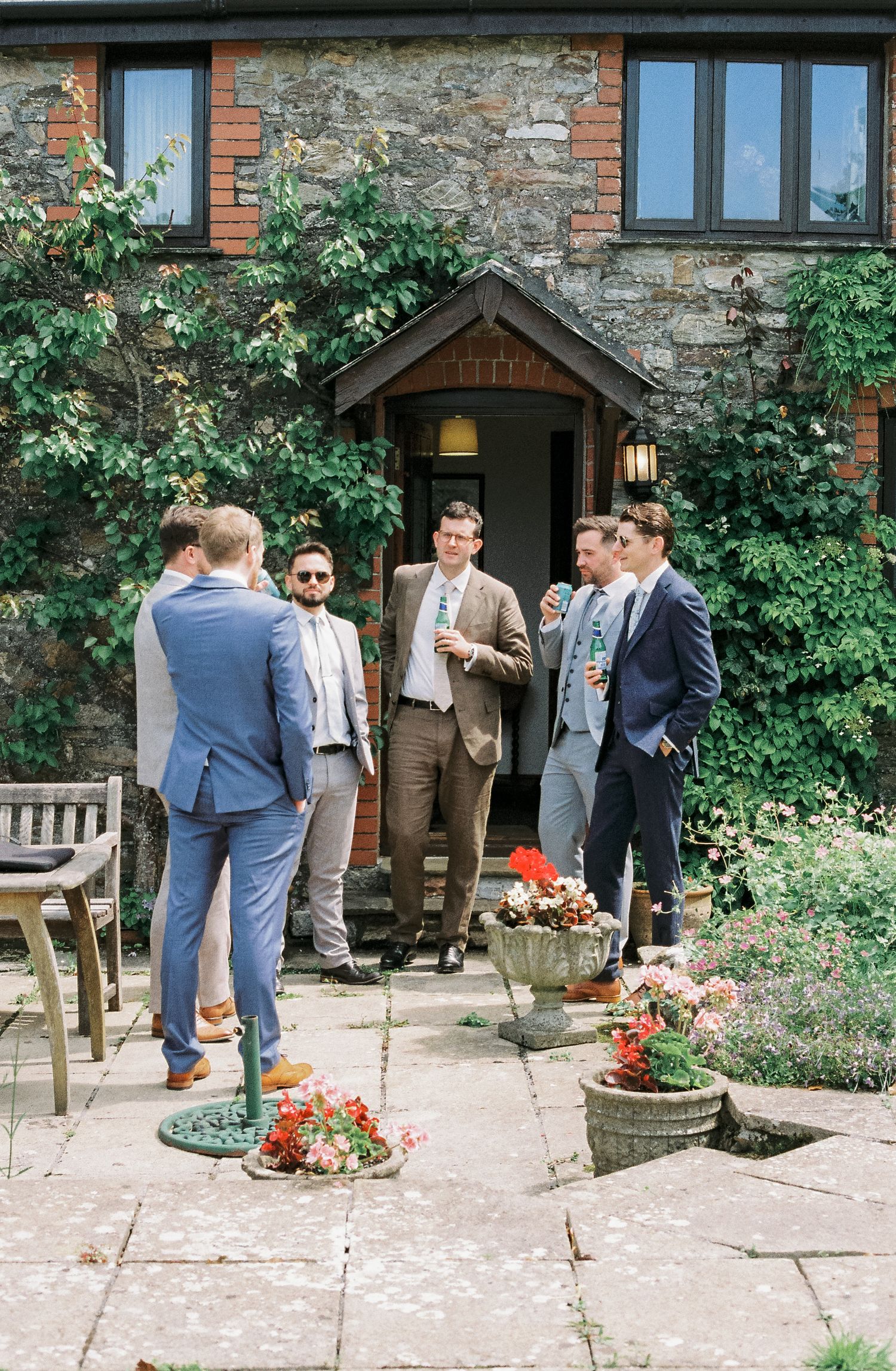 Wedding guests in formal attire gather outside a rustic brick and stone building.