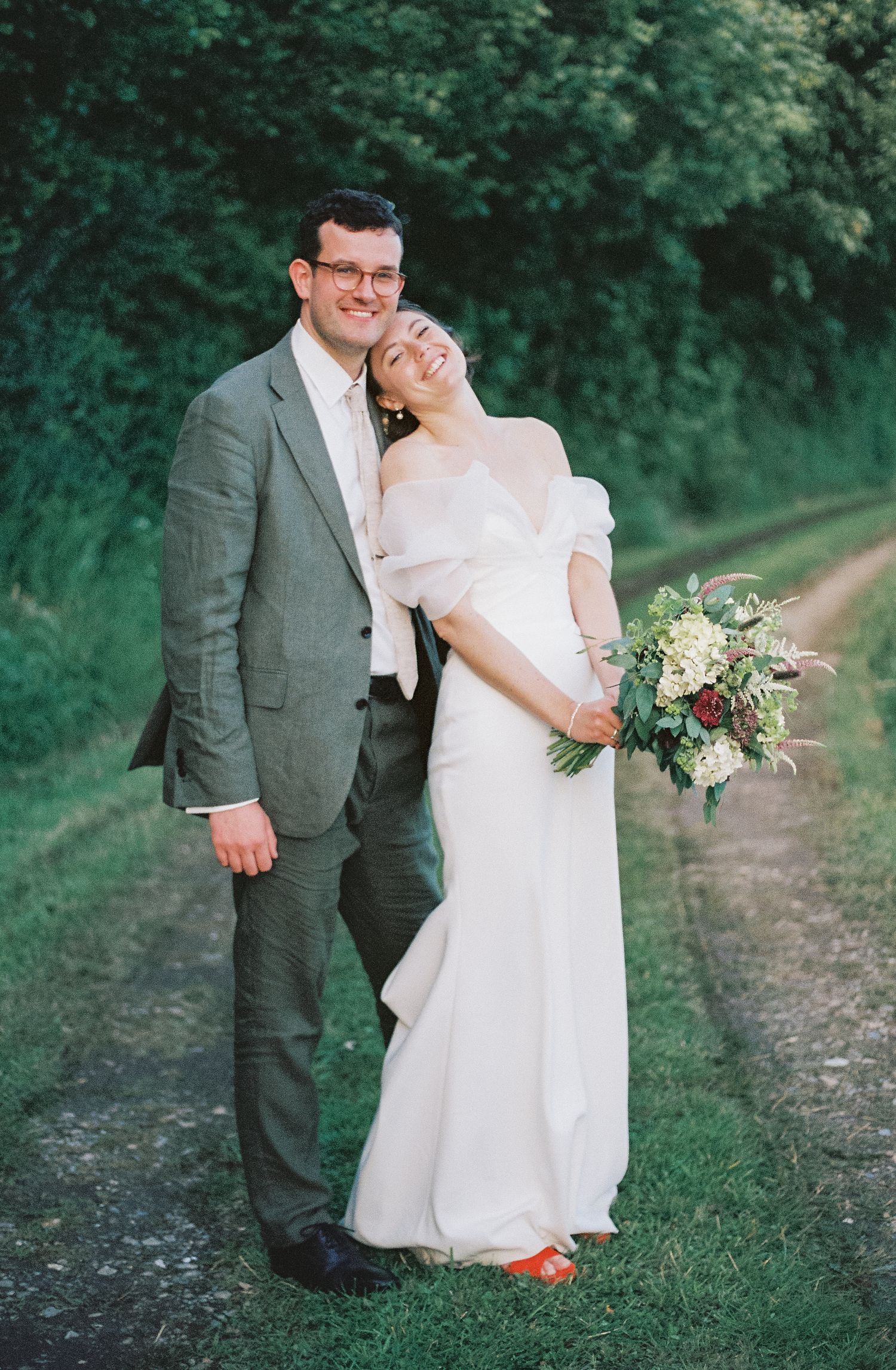 A bride in white dress and groom in grey suit share an intimate moment on a garden path.
