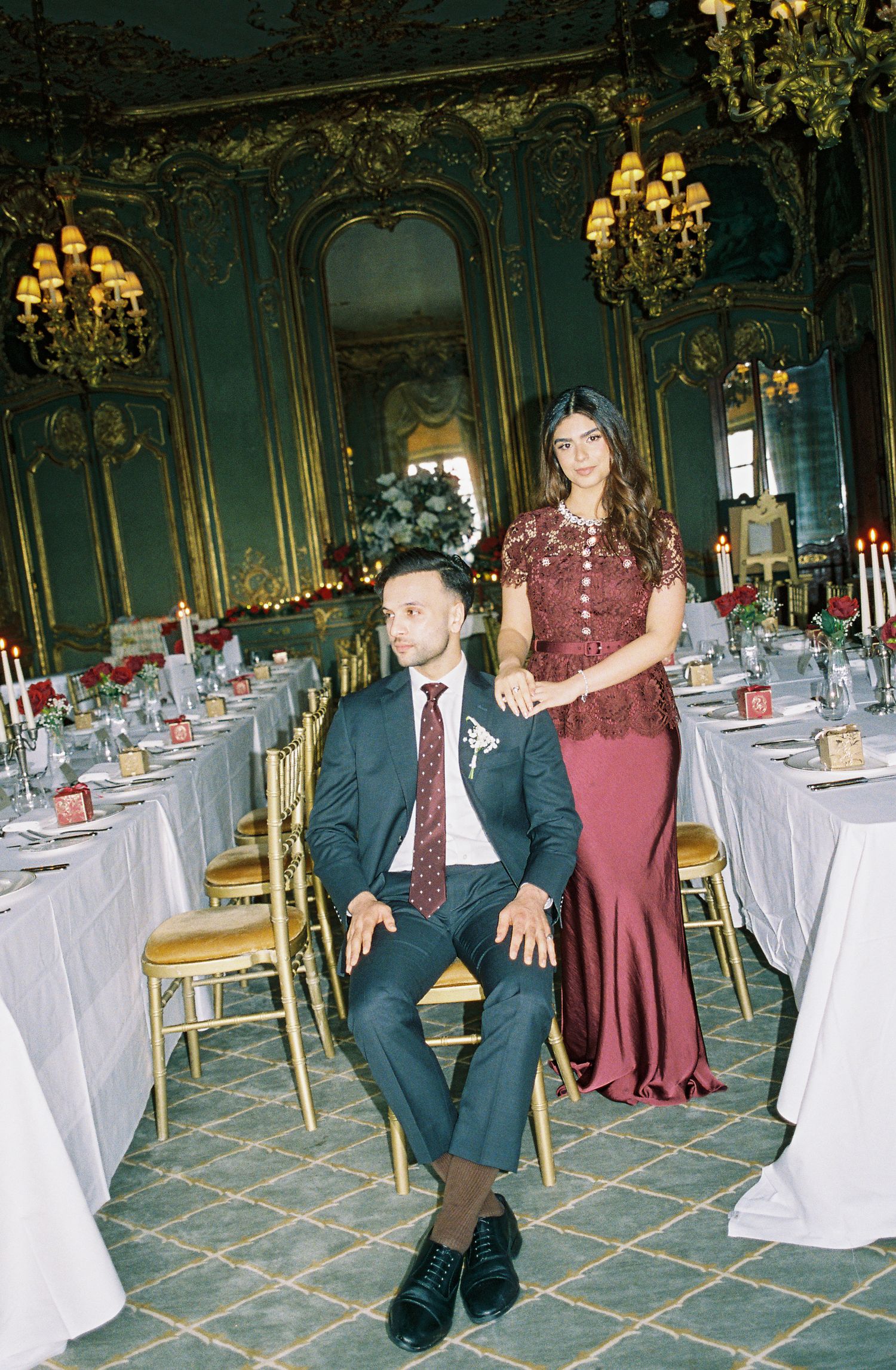 Elegant couple in burgundy and navy attire pose in ornate green ballroom with gold chandeliers and candlelit wedding tables.
