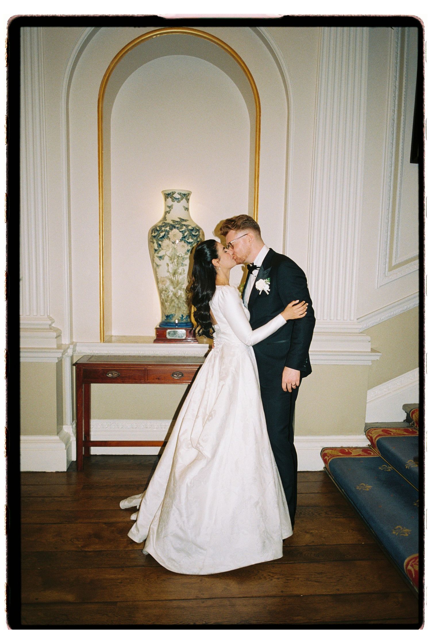Bride and groom share first kiss in elegant room, bride in long white gown, groom in black tuxedo.