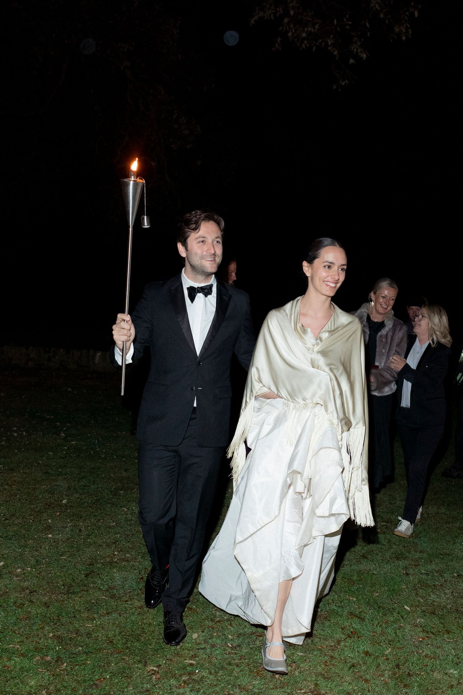 Groom in a black tuxedo carries a flaming torch beside a bride in a satin wrap as they walk across a lawn at night.