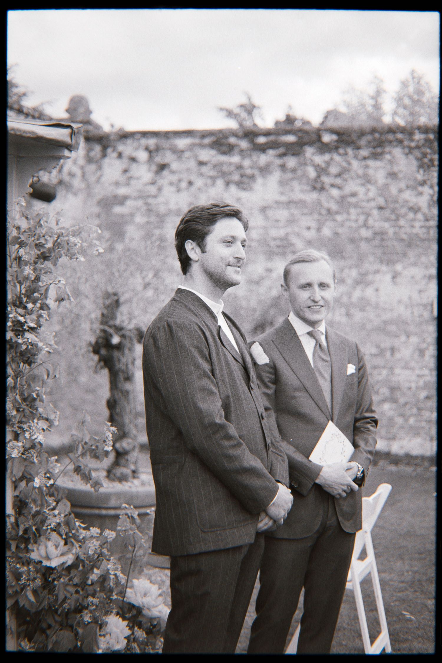 Two men in suits stand beside a weathered brick garden wall at an outdoor wedding ceremony, one holding a folded program.