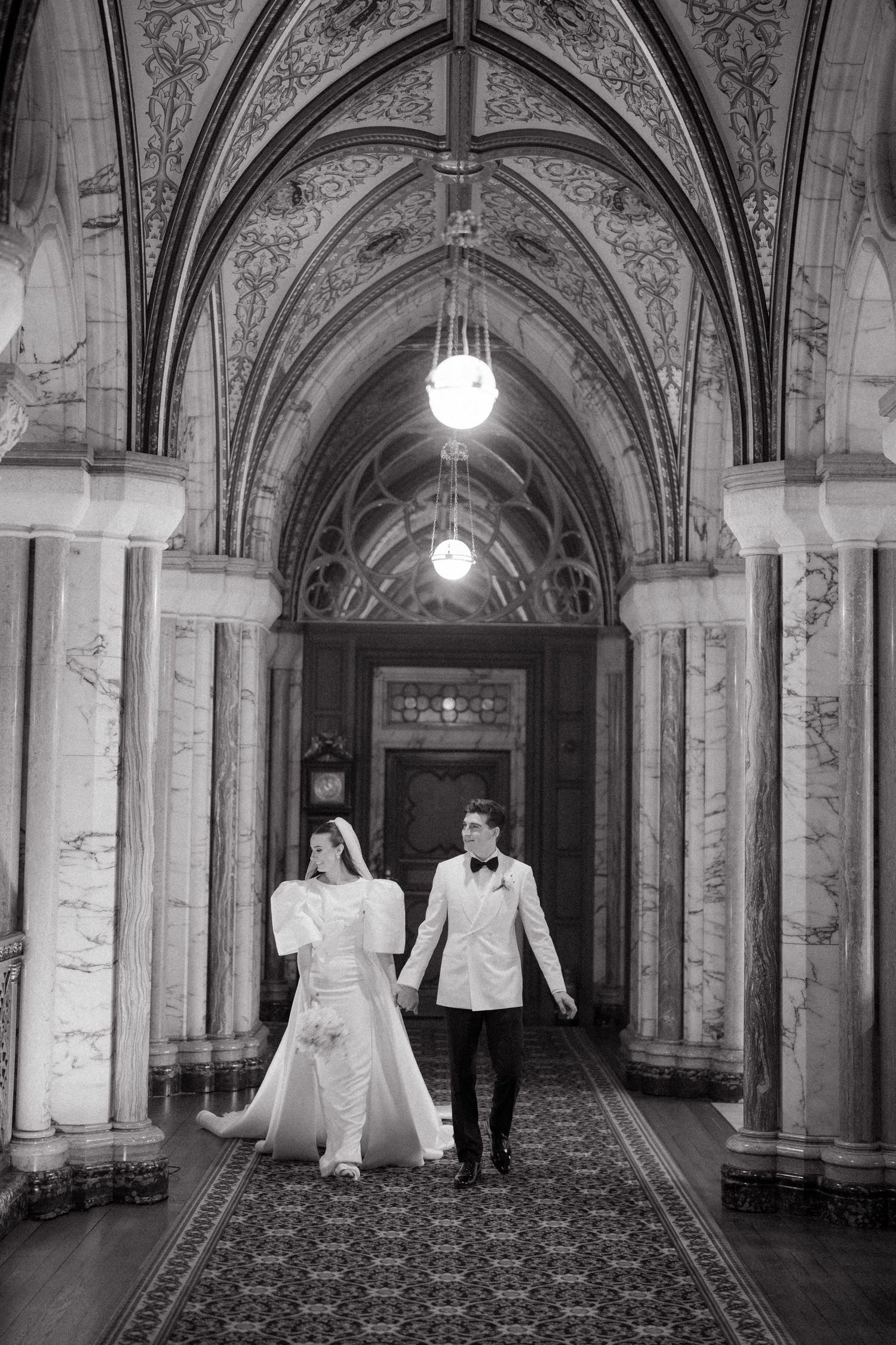 A black and white photo of a couple walking through a Gothic hallway with vaulted ceilings and ornate details.