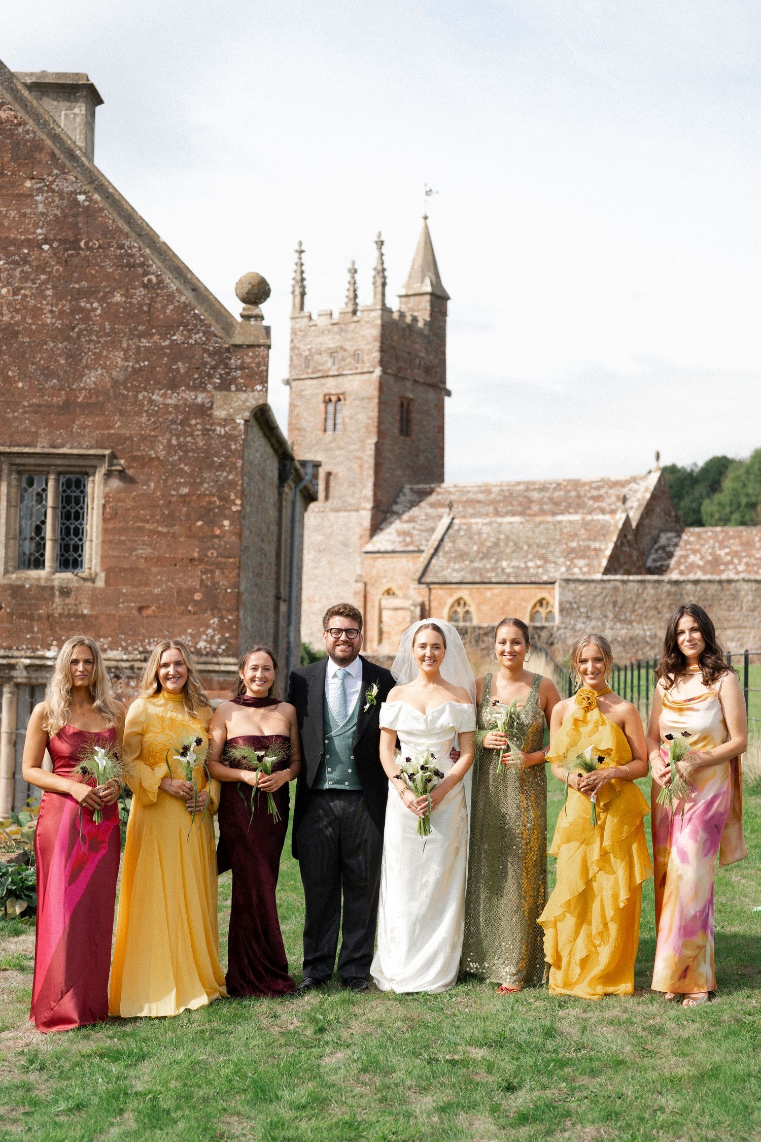 Wedding party poses outside historic church, bride in white gown with bridesmaids in colorful mismatched dresses holding bouquets.