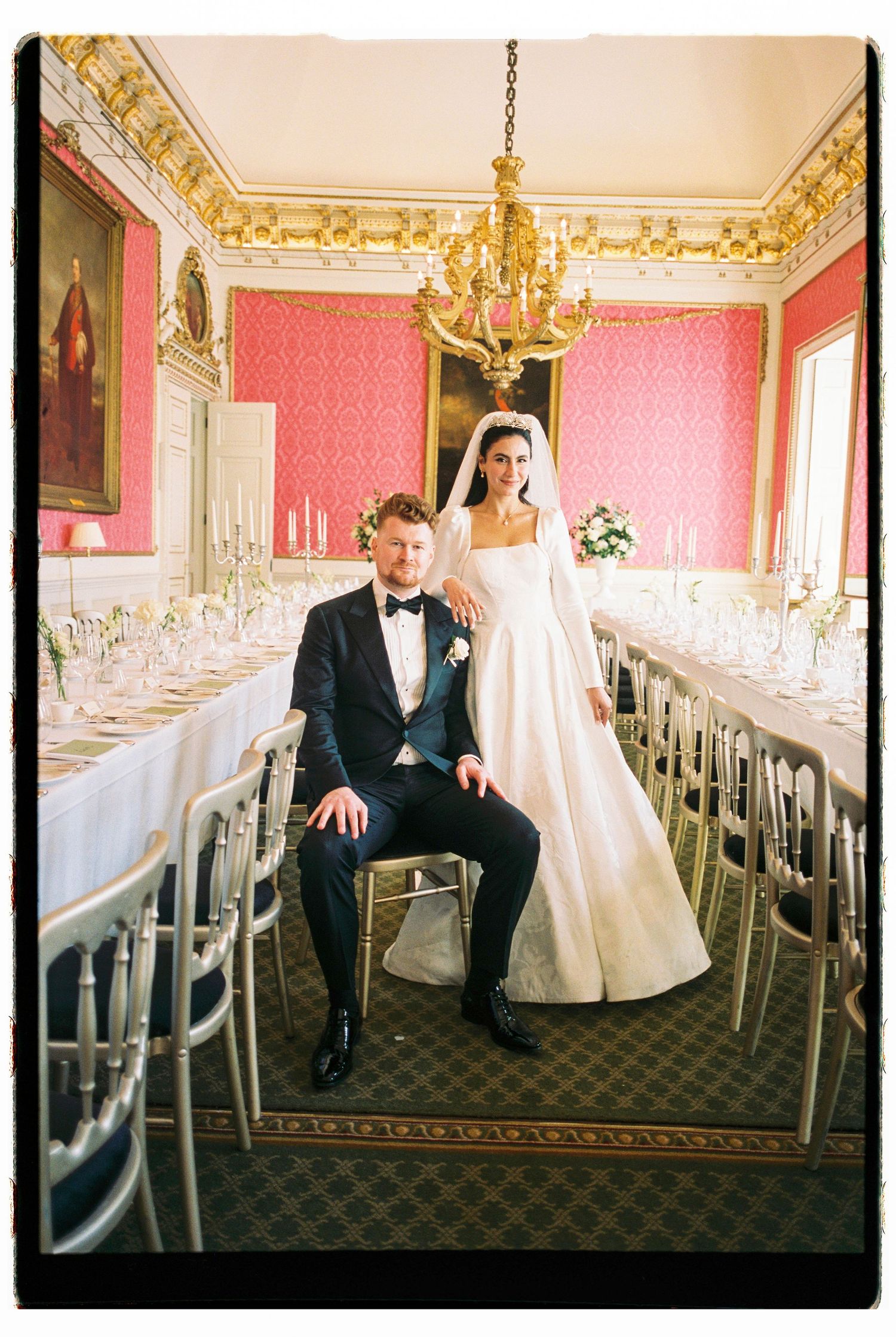 Bride and groom pose in ornate red dining room with gold chandelier and long reception tables.