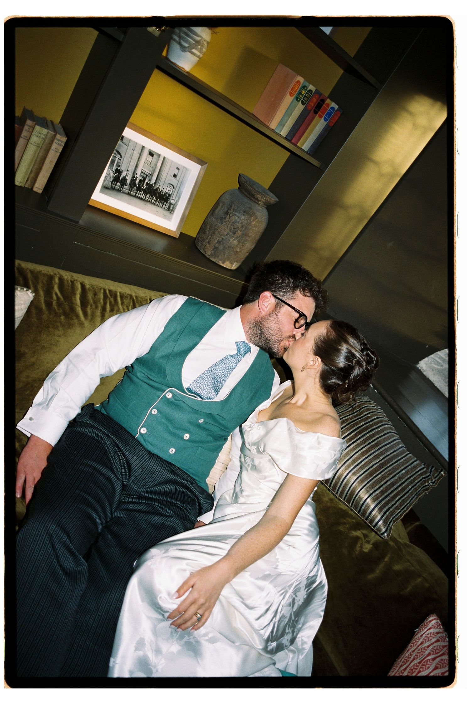 Newlywed couple shares a romantic kiss on a sofa, groom in teal waistcoat and bride in white gown.