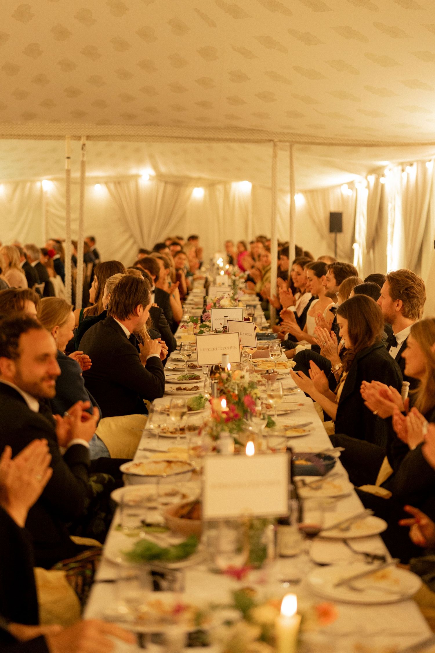 Wedding guests applauding along a long candlelit banquet table with flowers inside a softly lit white marquee.